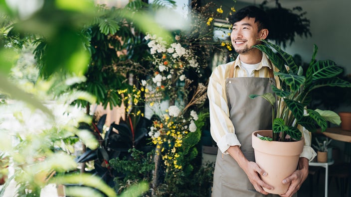 Plant shop owner carrying an indoor potted plant. He is smiling and surrounded by lush greenery.
