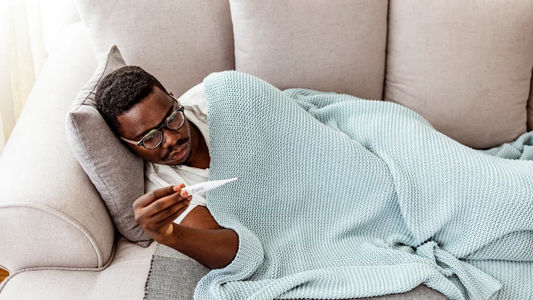Man with fever laying on couch at home with a crocheted blue blanket over him. He is reading the thermometer in his hand.
ljubaphoto/E+ via Getty Images