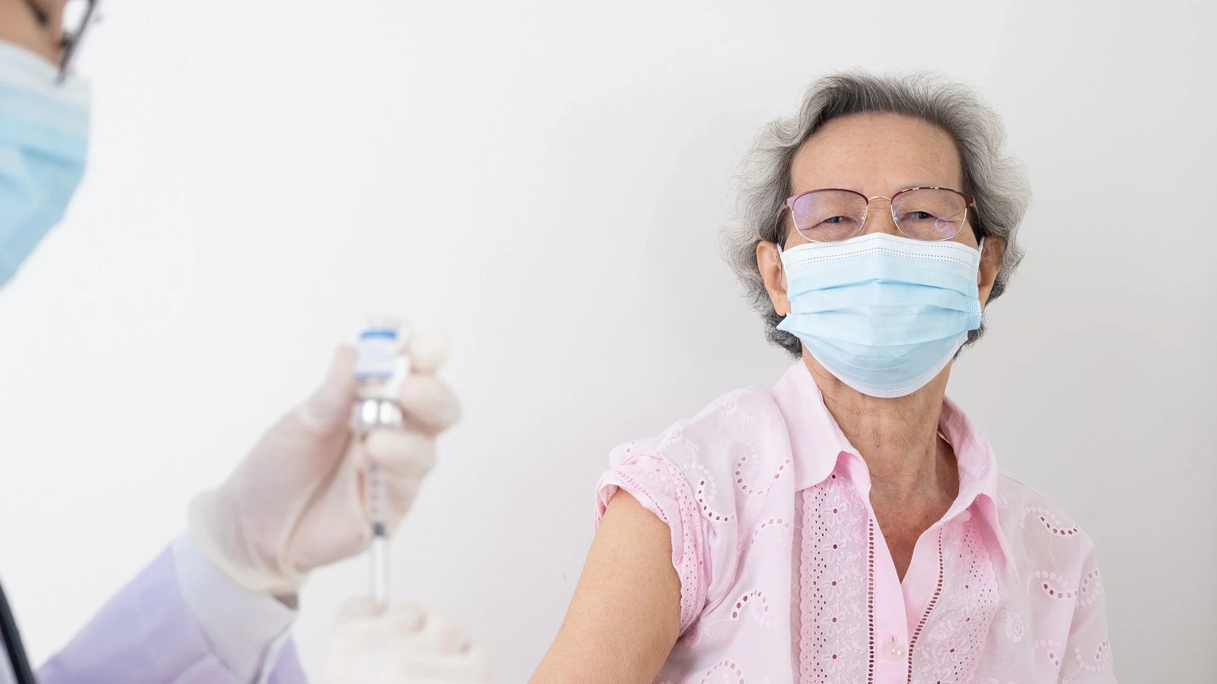 Portrait of an elderly woman getting the COVID vaccine. She is wearing glasses and a blue medical face mask. Her sleeve is rolled up ready for the shot while the nurse prepares the syringe.
Phawat via Shutterstock