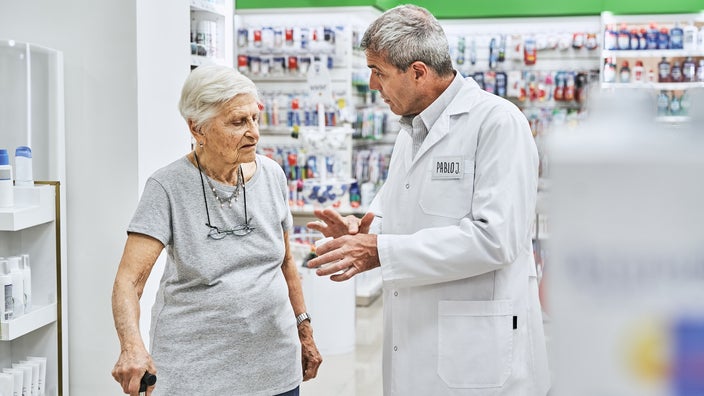 A senior woman speaks to a pharmacist inside a store.
