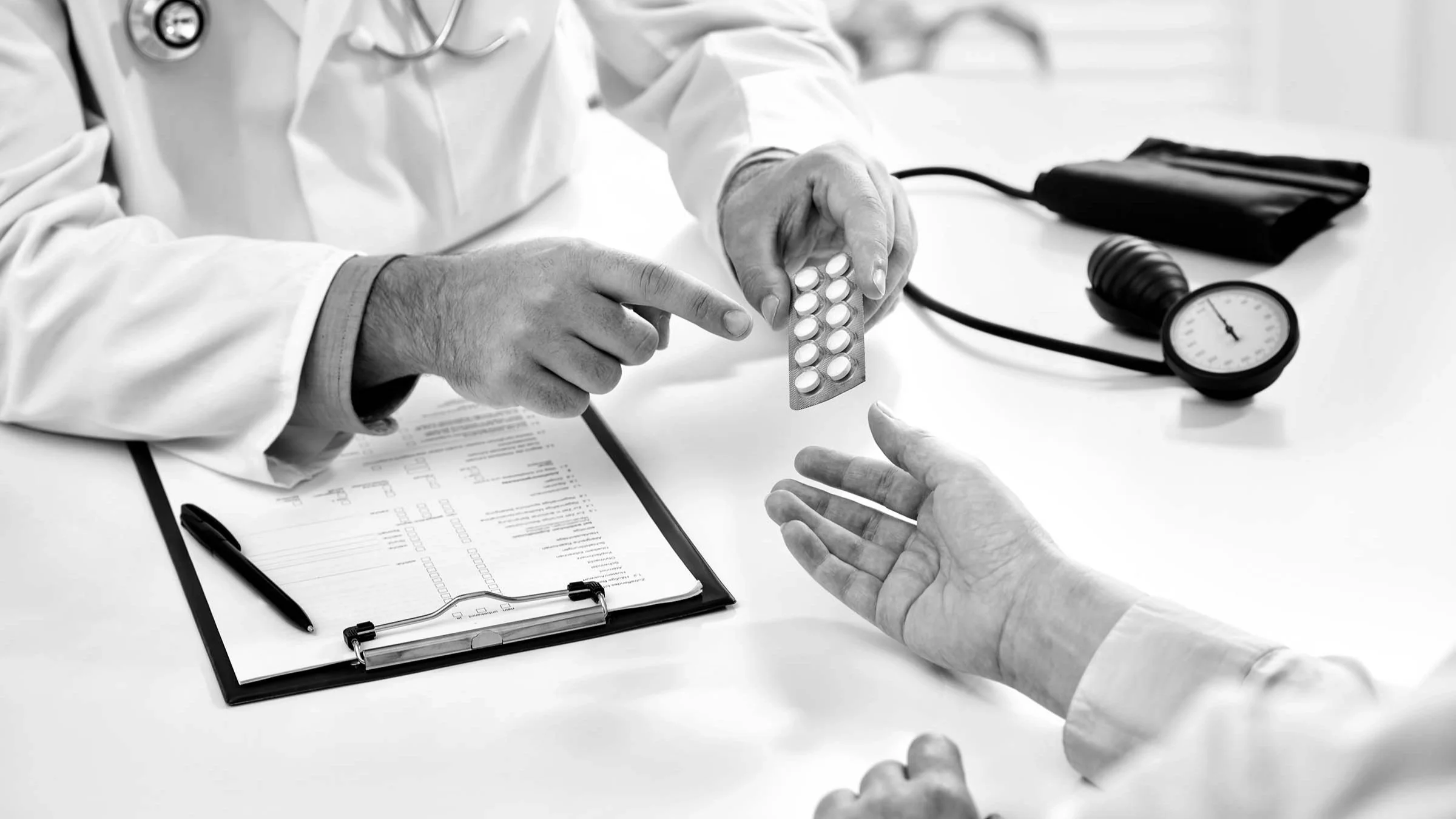 Black and white image of a doctor handing a patient a blister pack of medication across a desk.