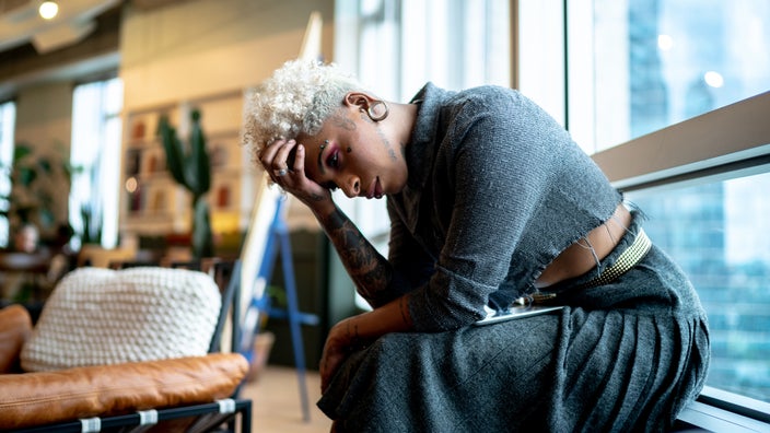 Young adult sitting off to the side at work with their head held down in their hand.