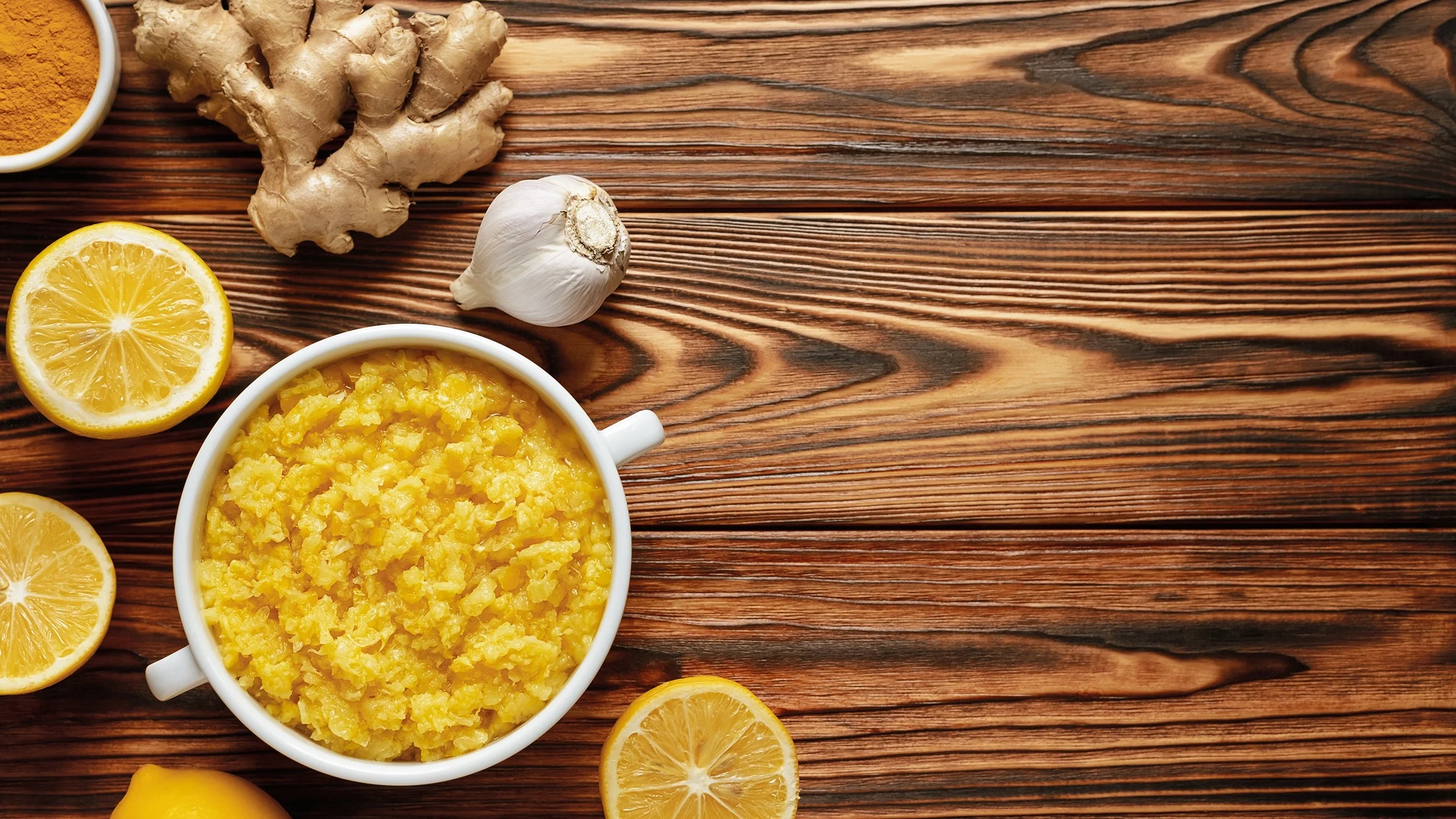 Bowl of garlic, ginger, turmeric, and lemon mixture on a wooden textured background. It is also surrounded by halved lemons, a bulb of garlic, and a head of ginger.