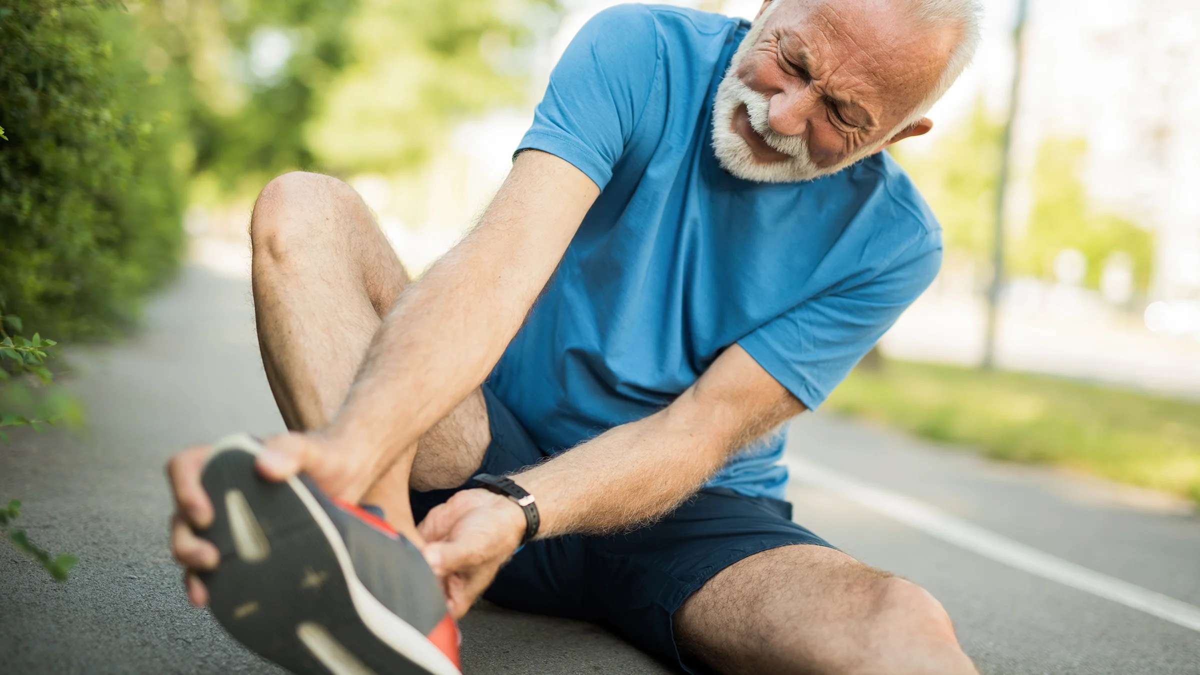 Senior man in the middle of a run stopping to see what is wrong with his foot. It looks like he is wincing in pain.
