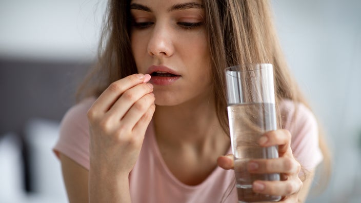 Close-up of a woman taking a pill with a glass of water. She is wearing a light pink t-shirt.