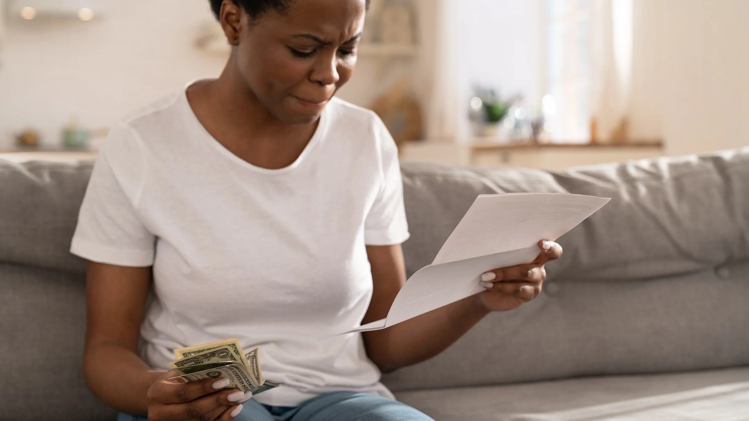 Young woman sitting on her couch holding a medical bill in one hand and cash in the other. She is pursing her lips together and looks upset.
