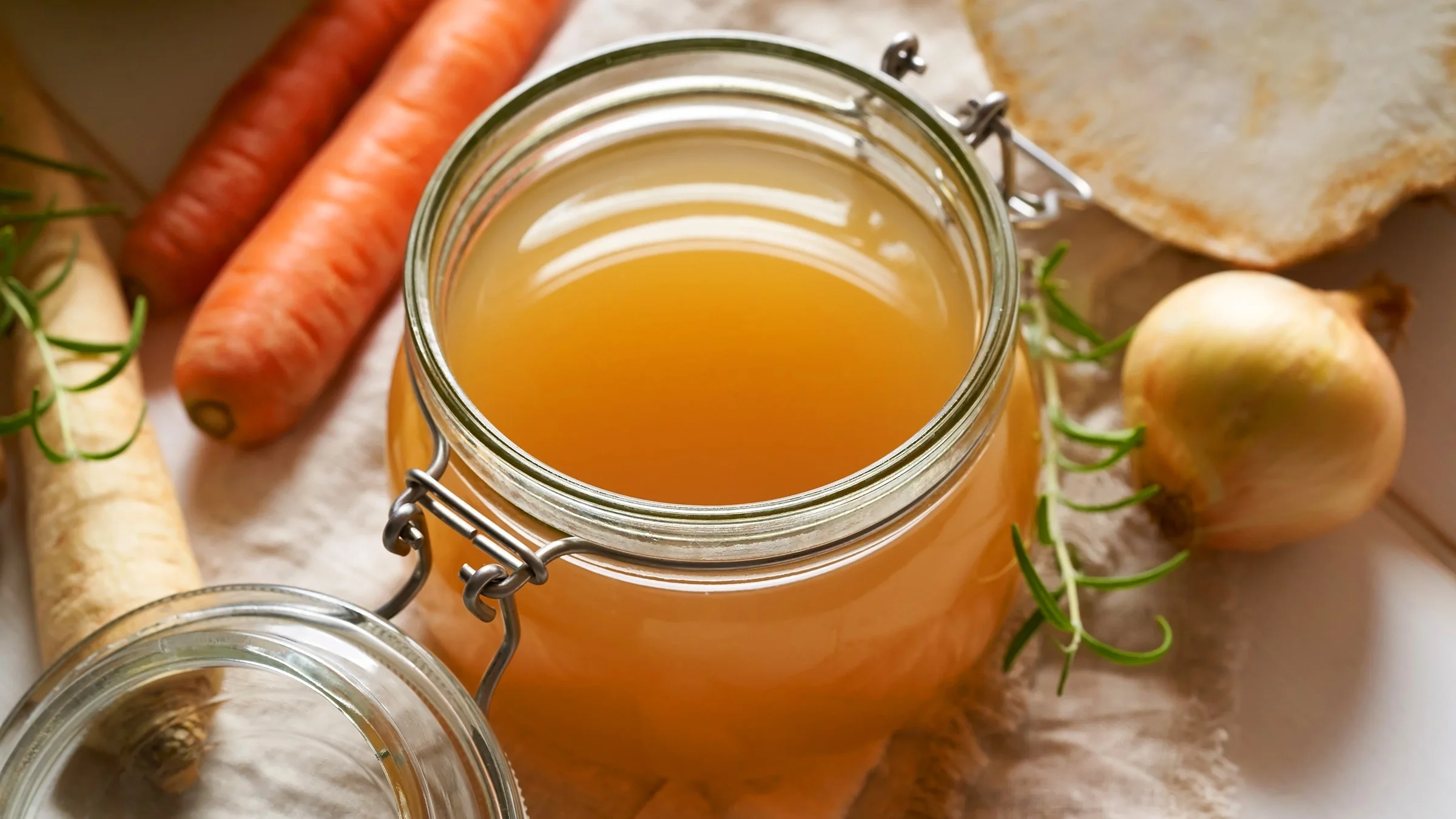 A homemade beef bone broth in a glass jar is pictured. Some carrots, onion, and other vegetables surround it.