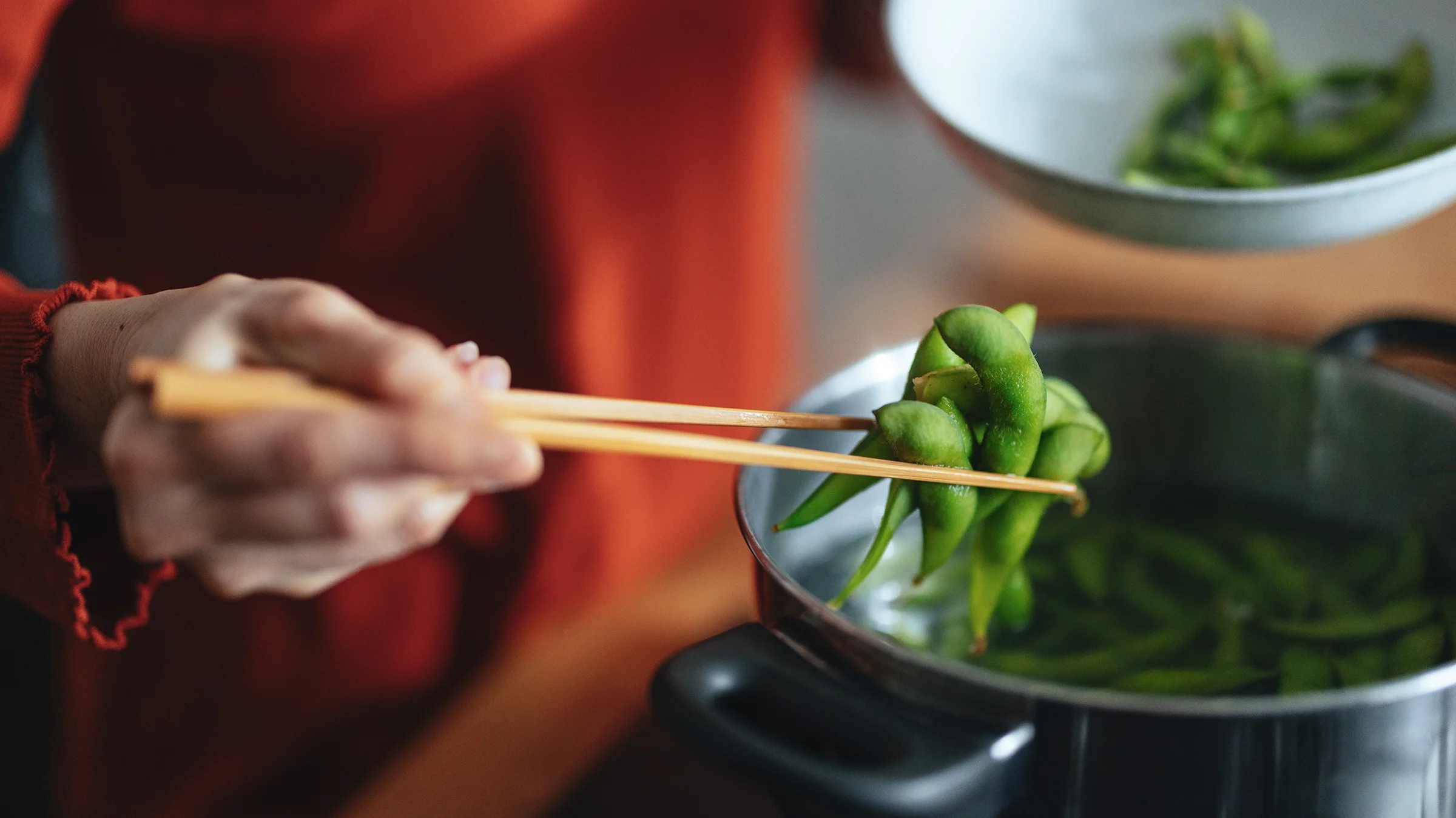 Close-up picking up cooked edamame beans with chopsticks from the pot.