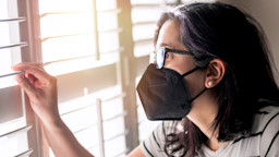 Young woman with a black face mask on looking out the window of her apartment.
Juanmonino/E+ via Getty Images