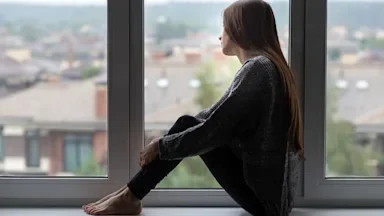 Side view of a young woman sitting on a window ledge indoors looking out the widow.
Yana Tikhonova/iStock via Getty Images