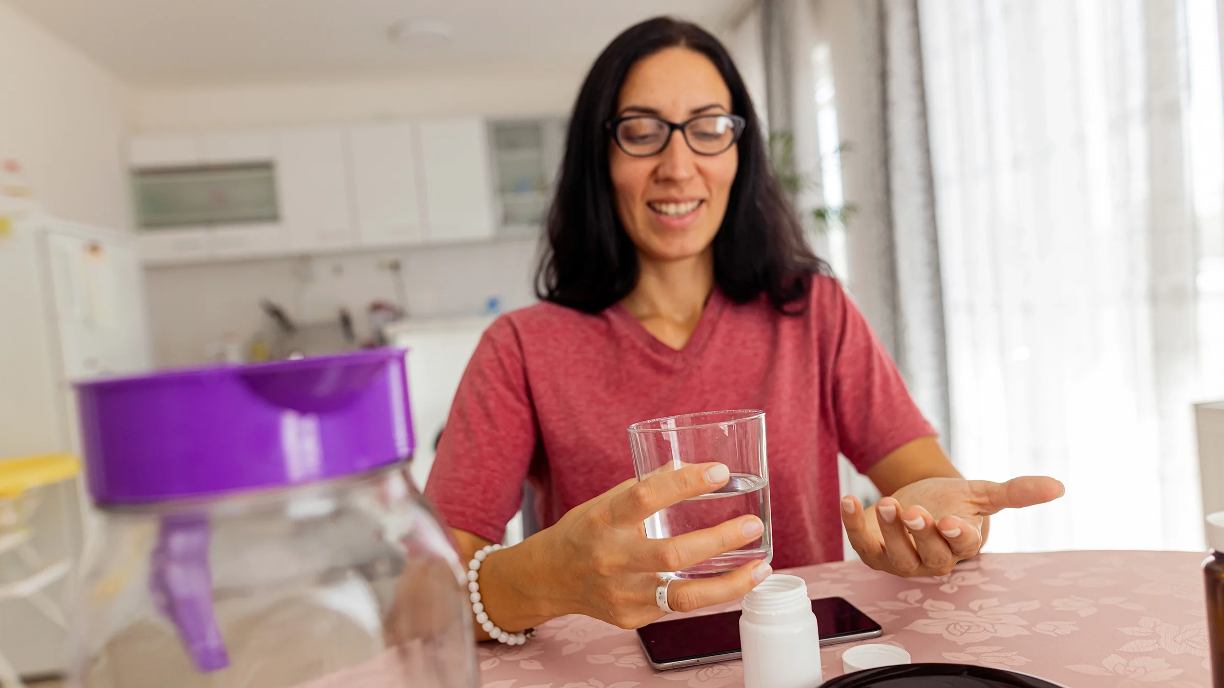 A woman sits at a dining table and takes medicine with a glass of water at home.