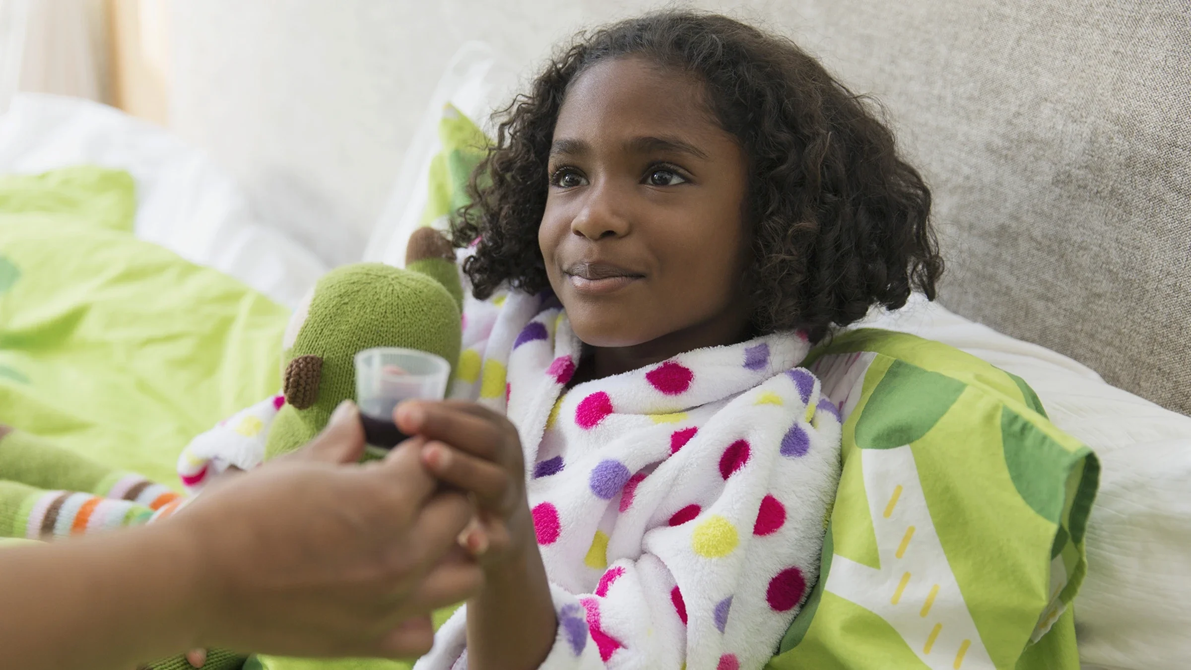A mother gives liquid medication to her daughter in bed.