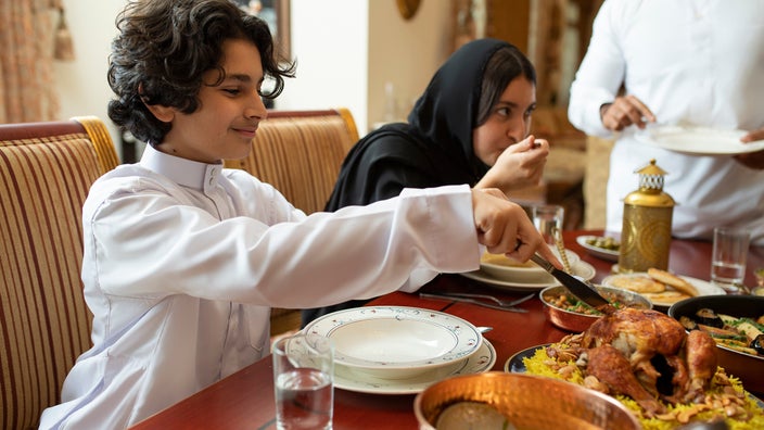Family enjoying a meal for Ramadan. The focus is on a child serving food and women in the background wearing a black Hijab.