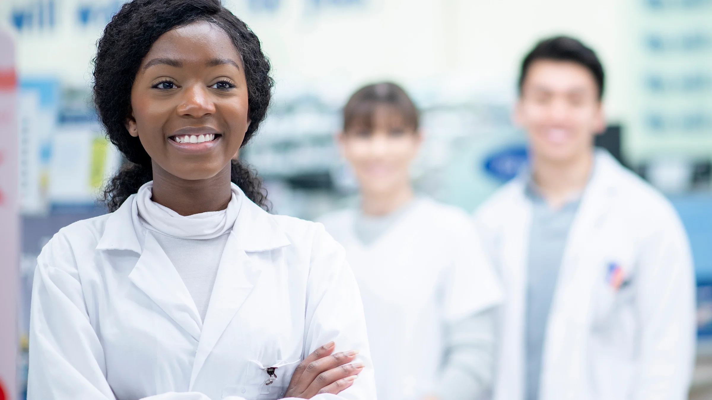A pharmacist stands with her arms crossed, and two other pharmacists stand in the background.