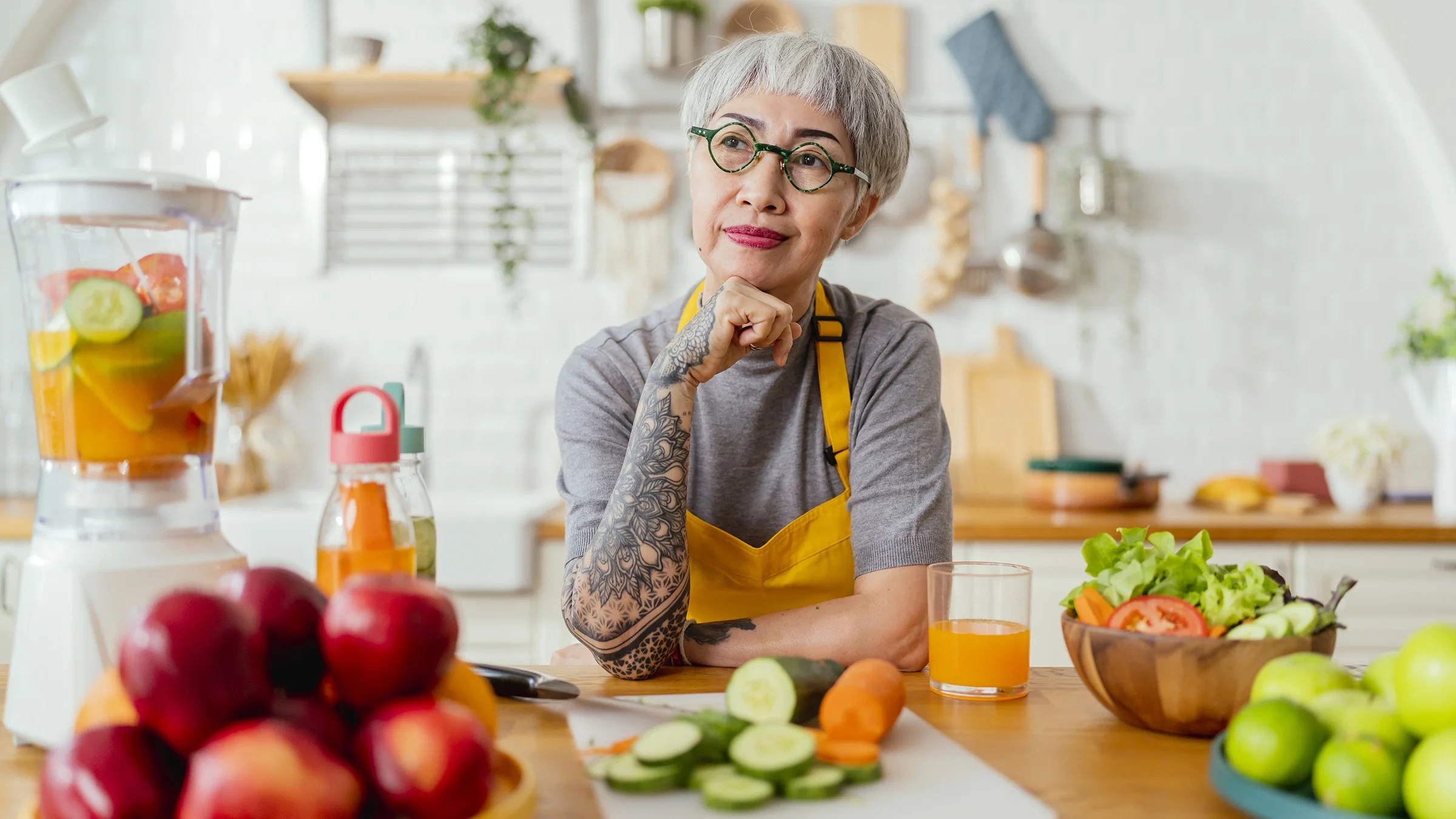A woman sits in a kitchen with a chopped carrot and cucumber in front of her.
