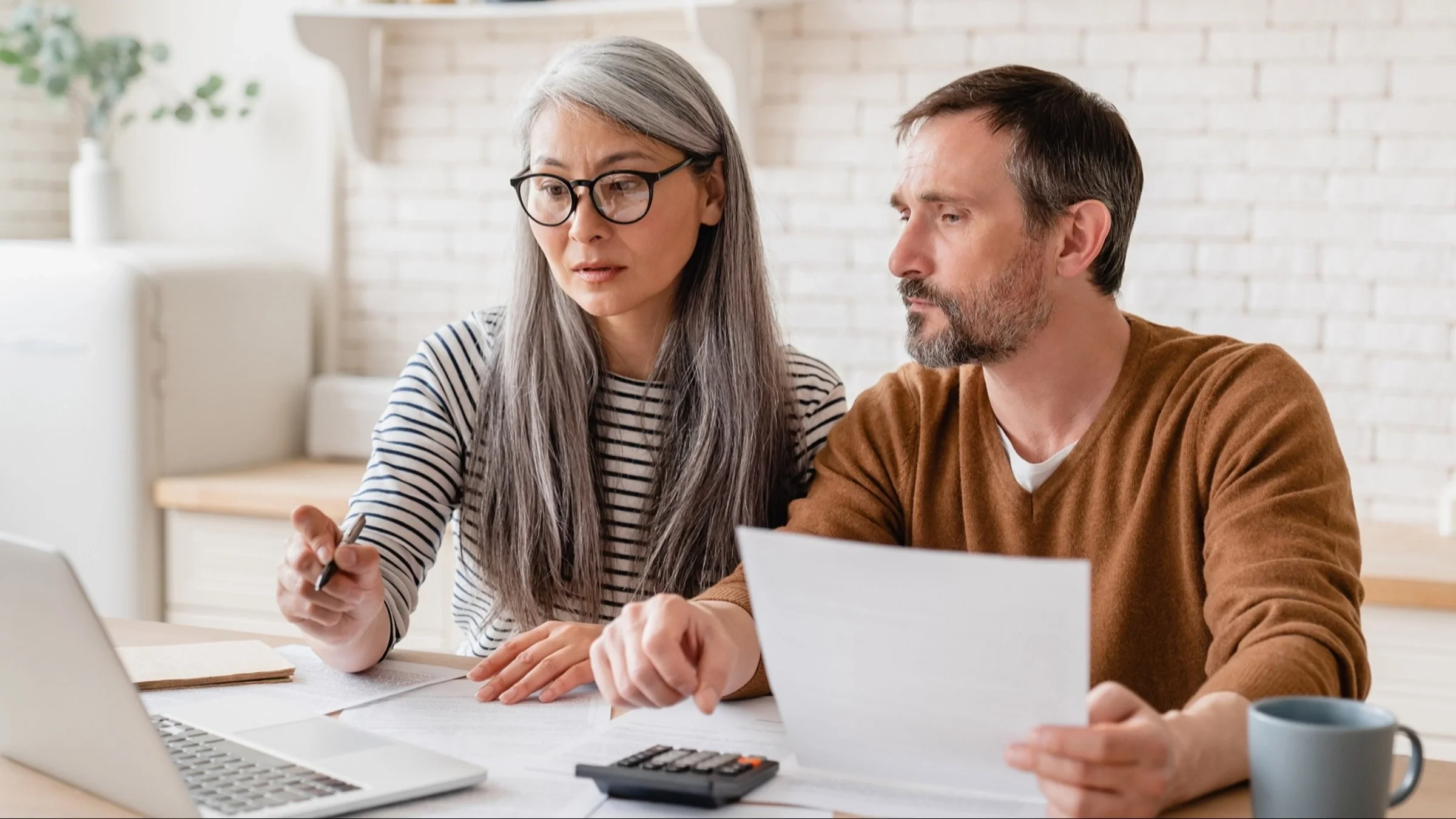 Mature older couple sitting at the kitchen table going over bills with their laptop open. They both look really focused on something on the laptop screen.