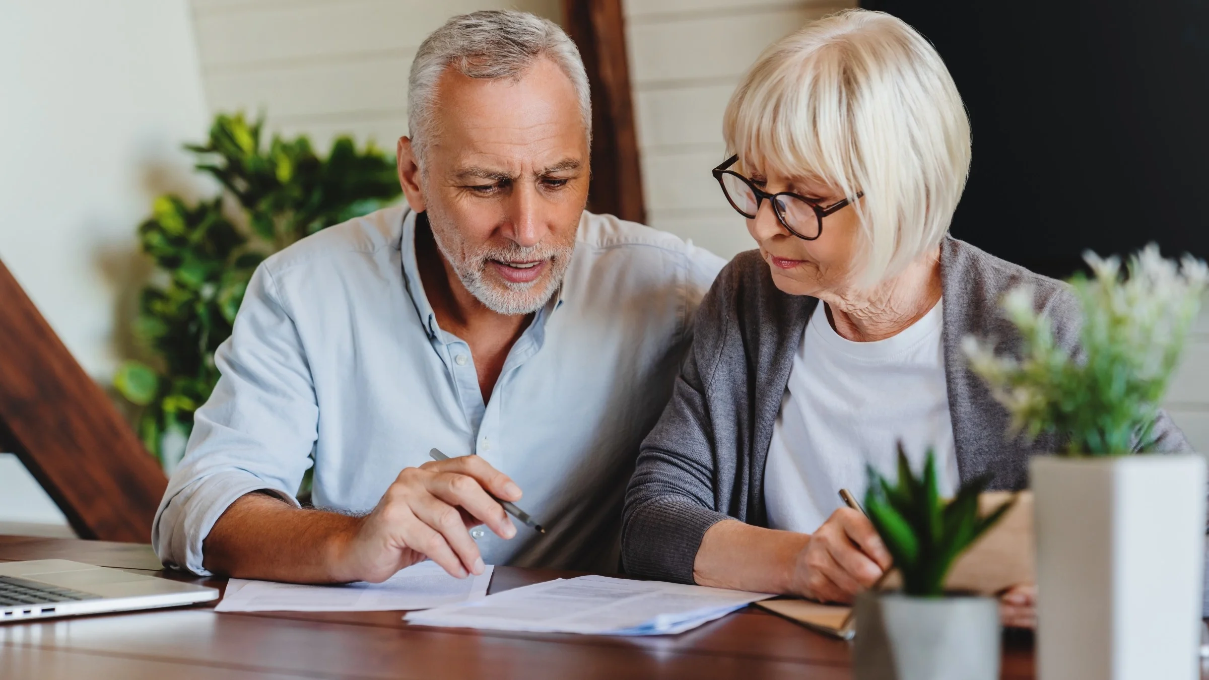 Older couple reviewing paperwork and documents at their dinning table with different house plants surrounding them.