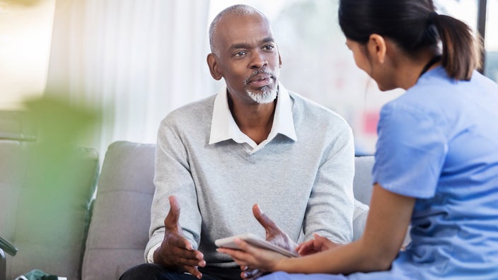 Senior man speaking with his doctor. He is sitting on a couch leaning to talk to the doctor who is on a chair next to him.