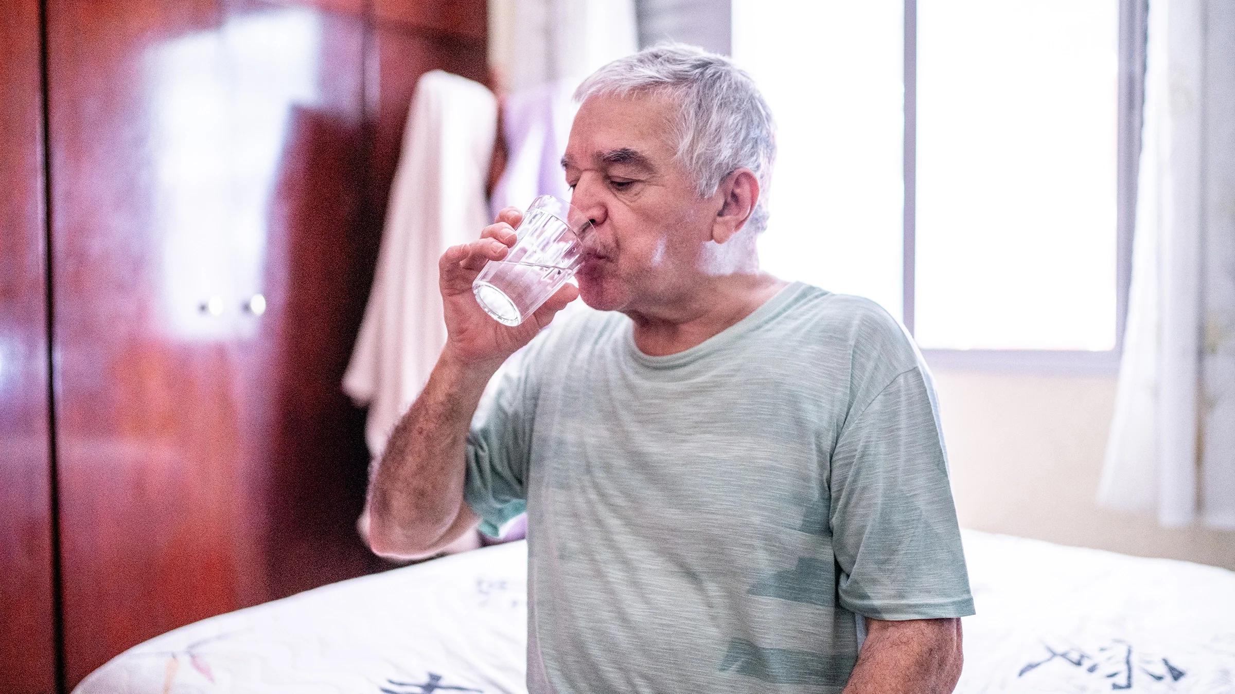 A man drinks a glass of water in his bedroom.