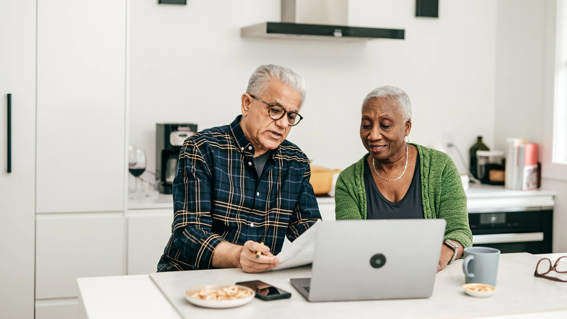 A senior couple are sitting in their kitchen. They are looking at banking paper statements together.