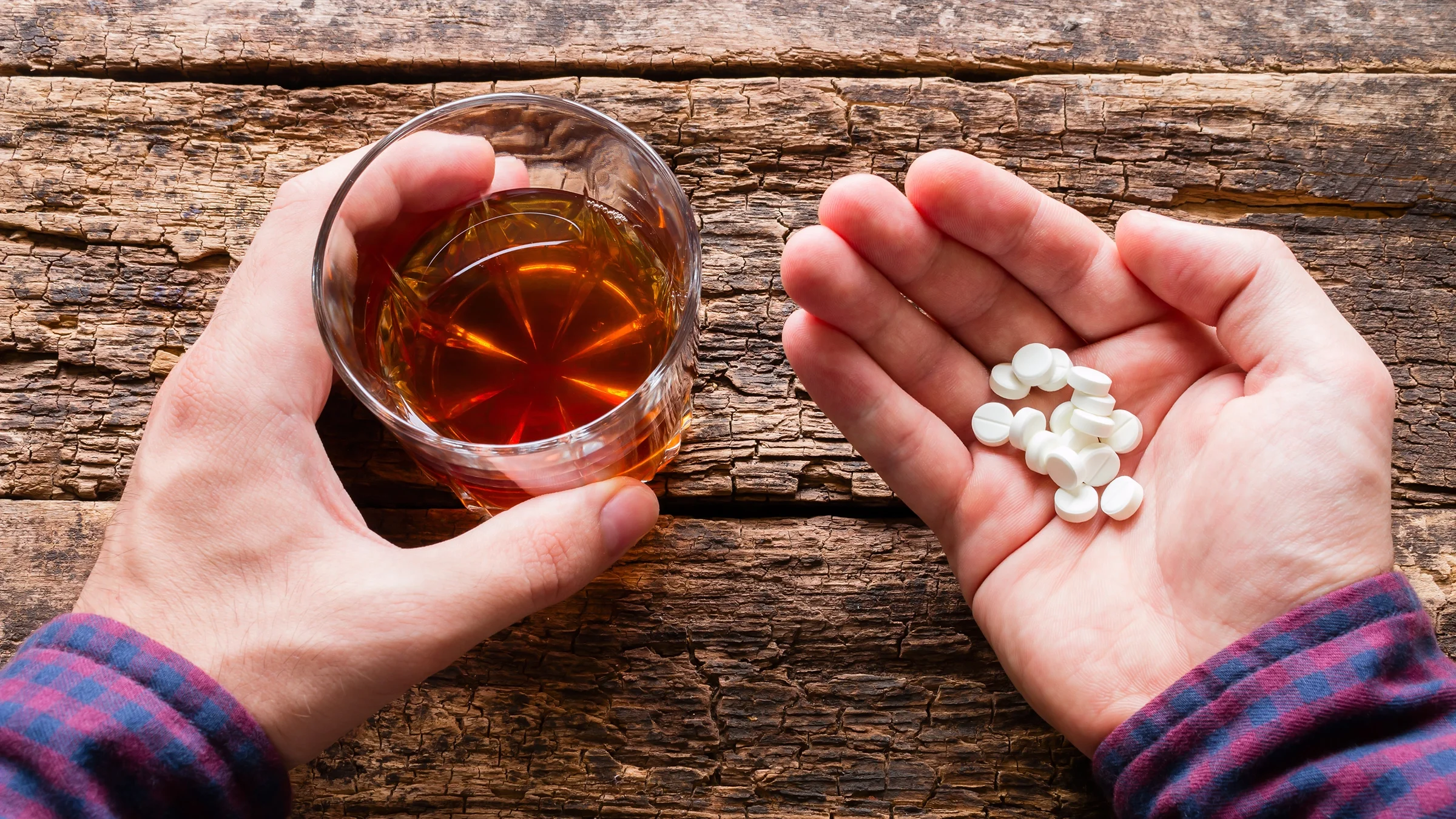 Close-up of hands holding pills and a glass of whiskey on a wooden background.