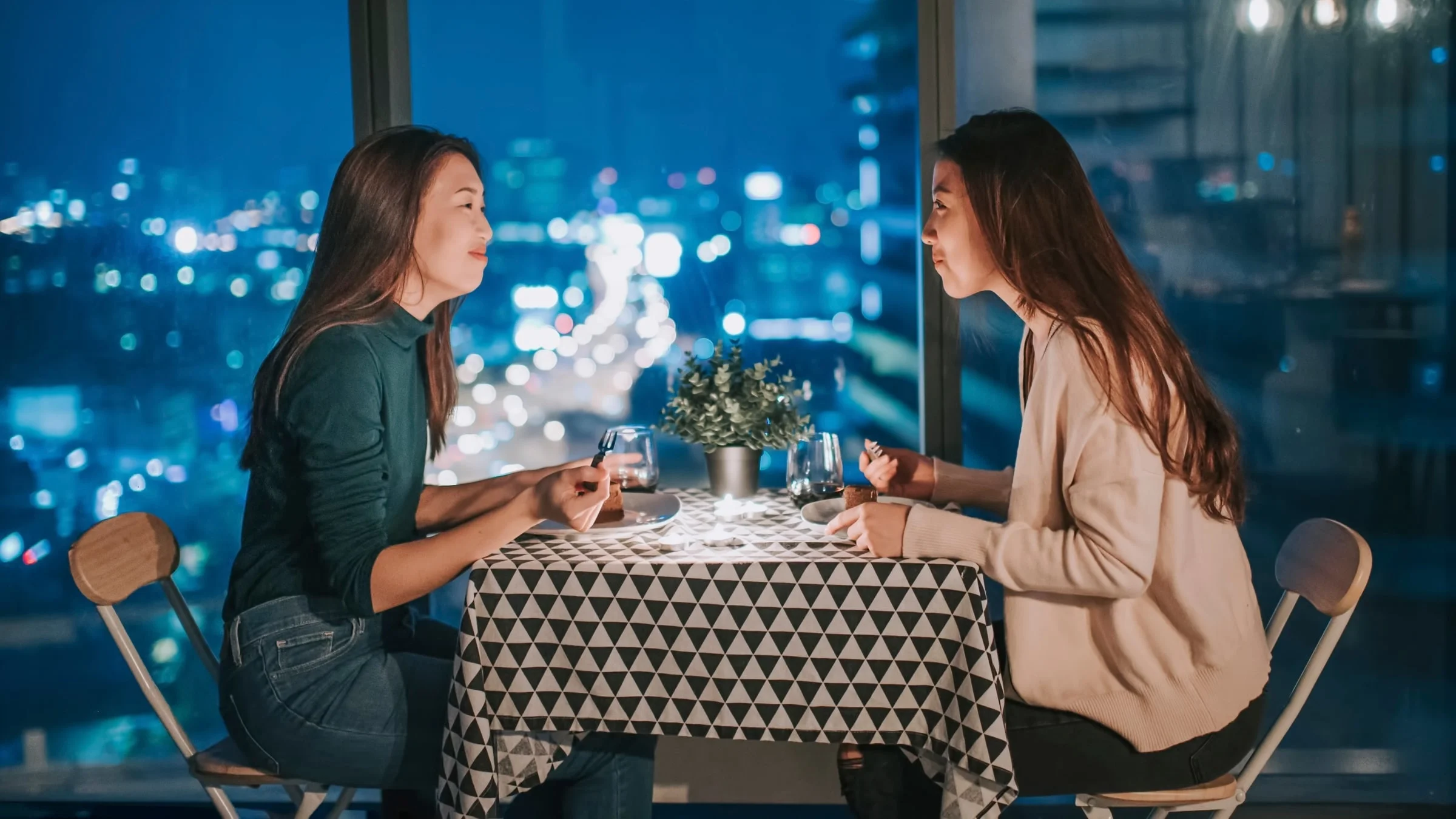 A lesbian couple talking over dinner at a restaurant with a city view.