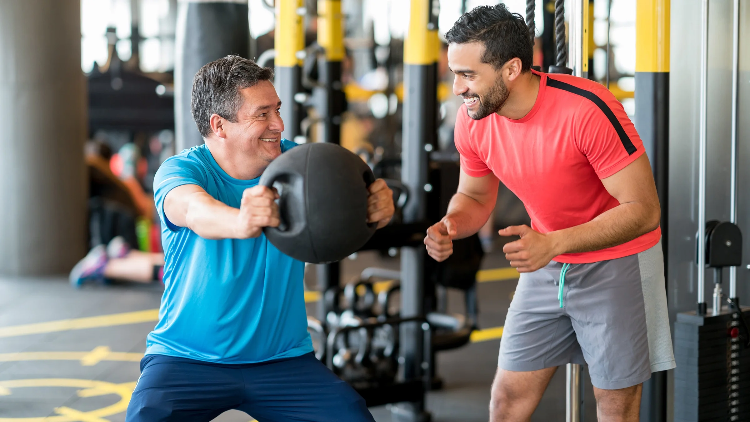 Middle aged man working out with his trainer at the gym. He is doing squats with a weighted ball.