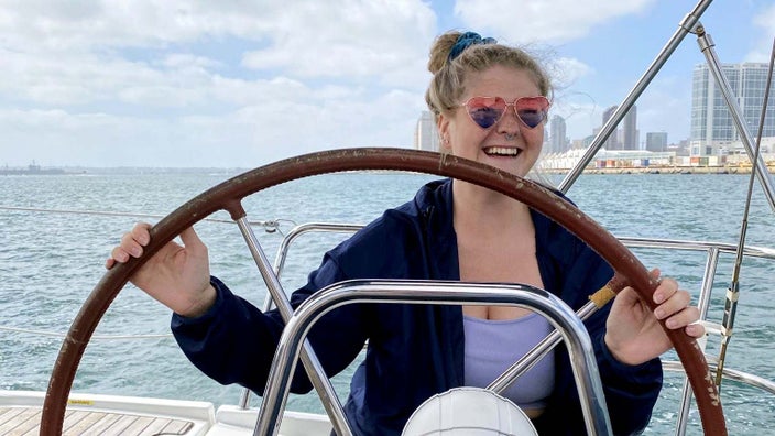 Hayley Chamblin holding a nautical wheel by the water. She is wearing heart-shaped sunglasses.