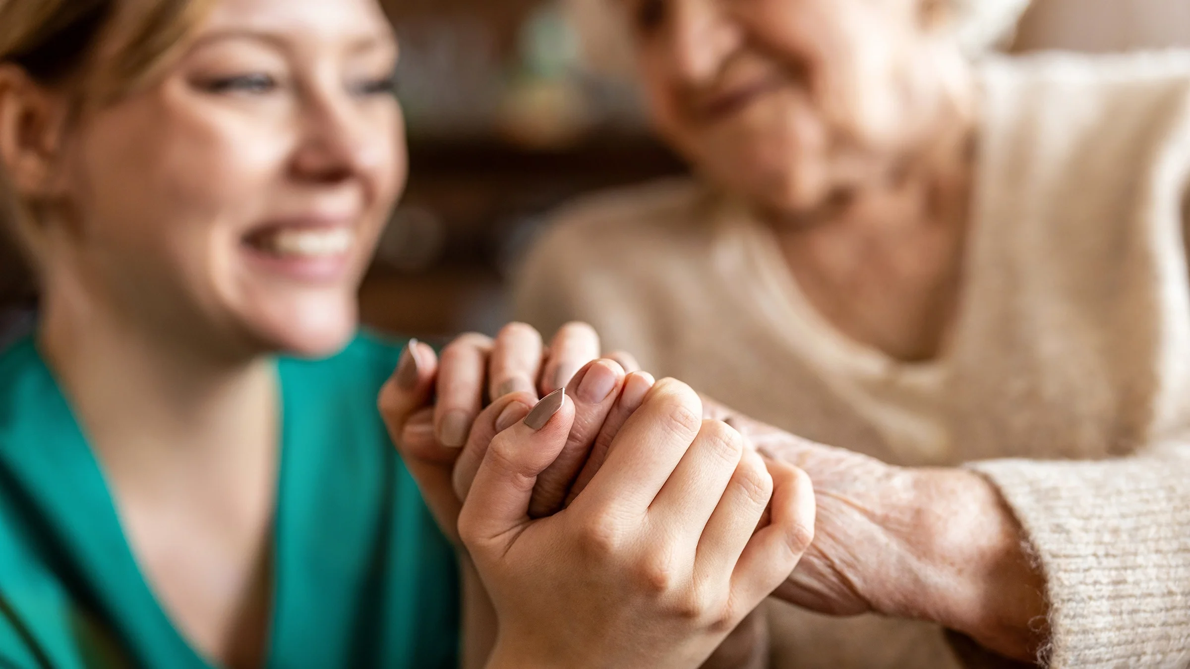 Close-up on a nurse holding her patient's hands. You can see them in the blurry background smiling.