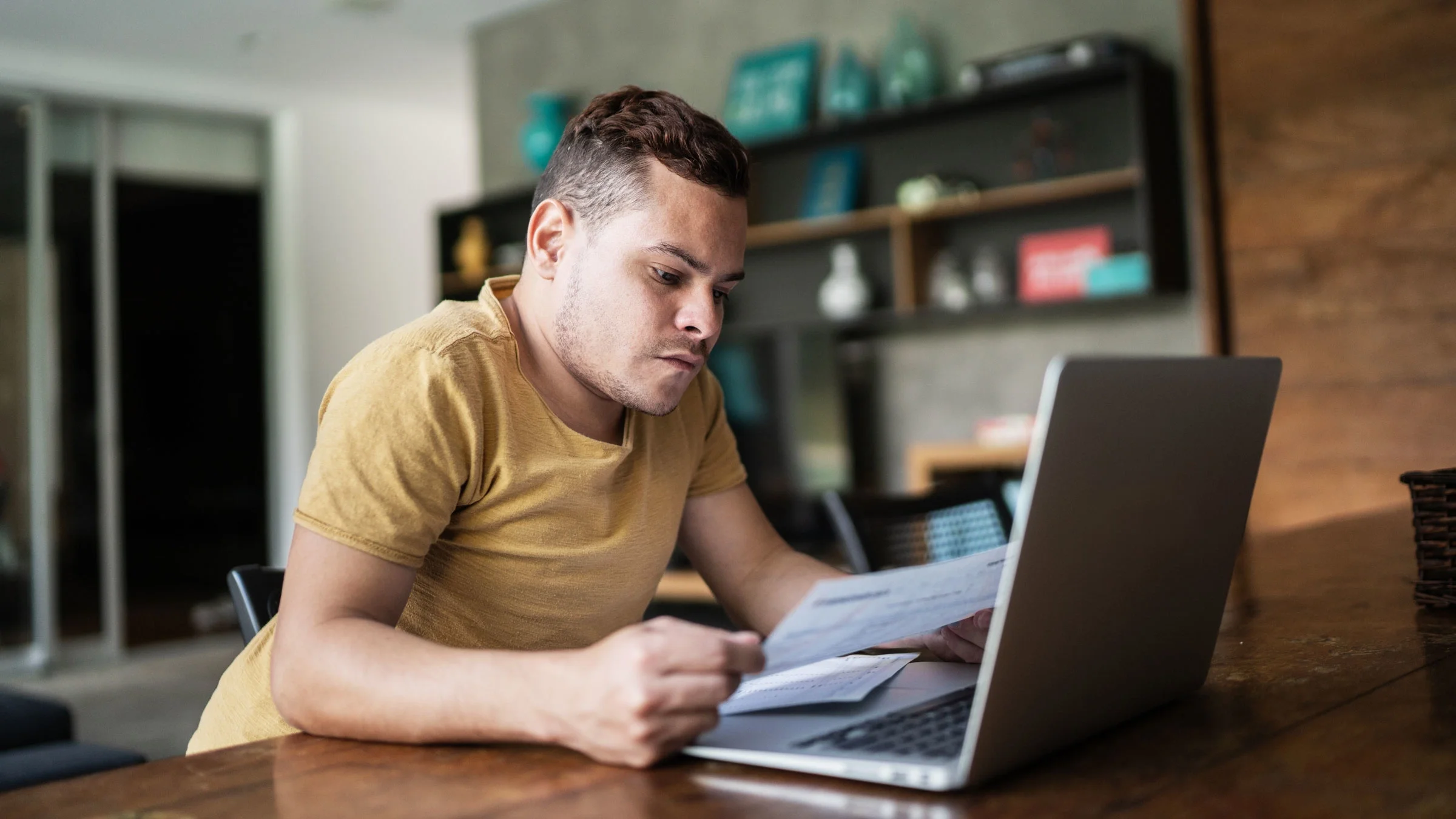 A man is looking at financial paperwork, leaning over a laptop.