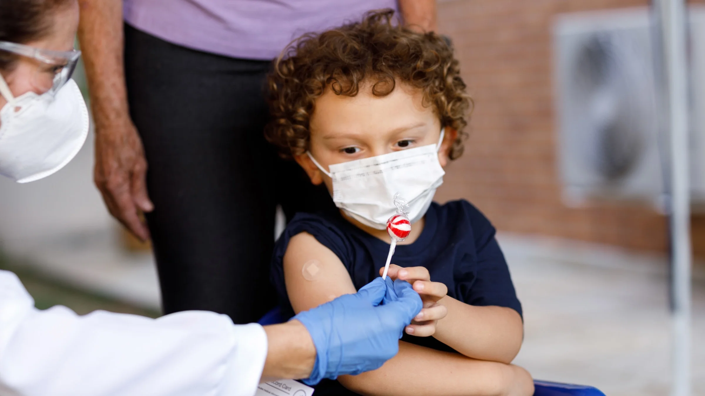 A little boy getting a lollipop from a healthcare provider, after getting a vaccine. There's a bandaid on his arm.