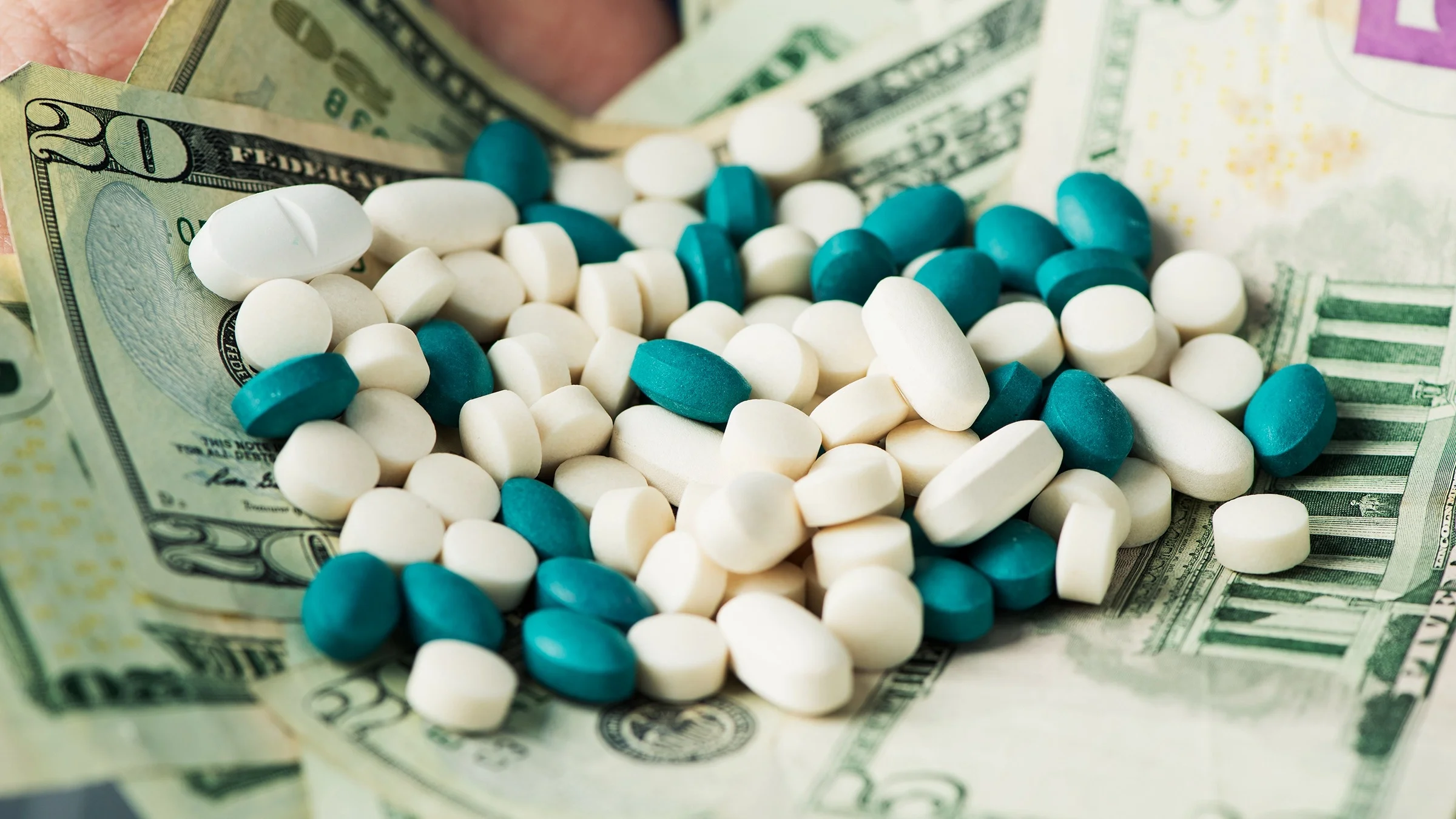 Various white and blue/green pills on a pile of money. You can see a little bit of a person's hand in the background holding it all.
