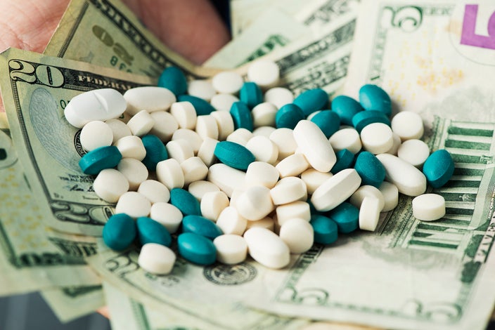 Various white and blue/green pills on a pile of money. You can see a little bit of a person's hand in the background holding it all.