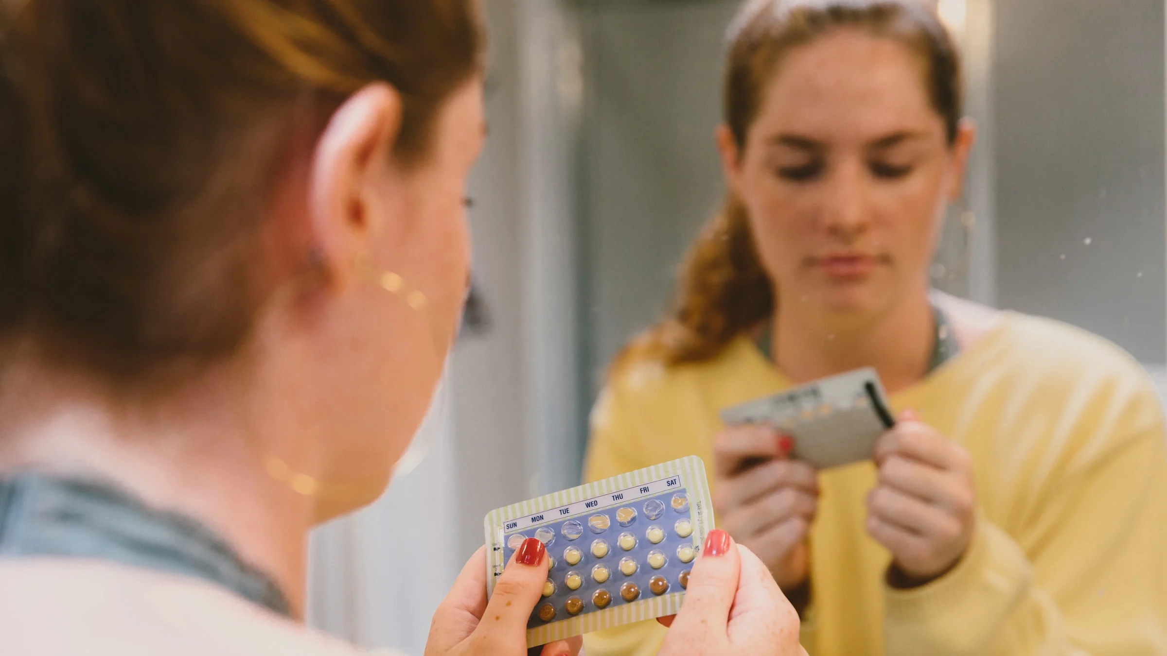 A person holding a birth control packet, standing in front of their bathroom mirror.