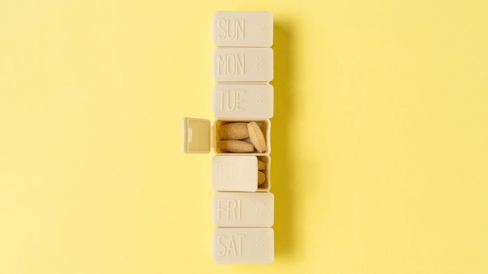 Tan pill organizer on a light yellow background one of the days of the week is open to show vitamins inside.