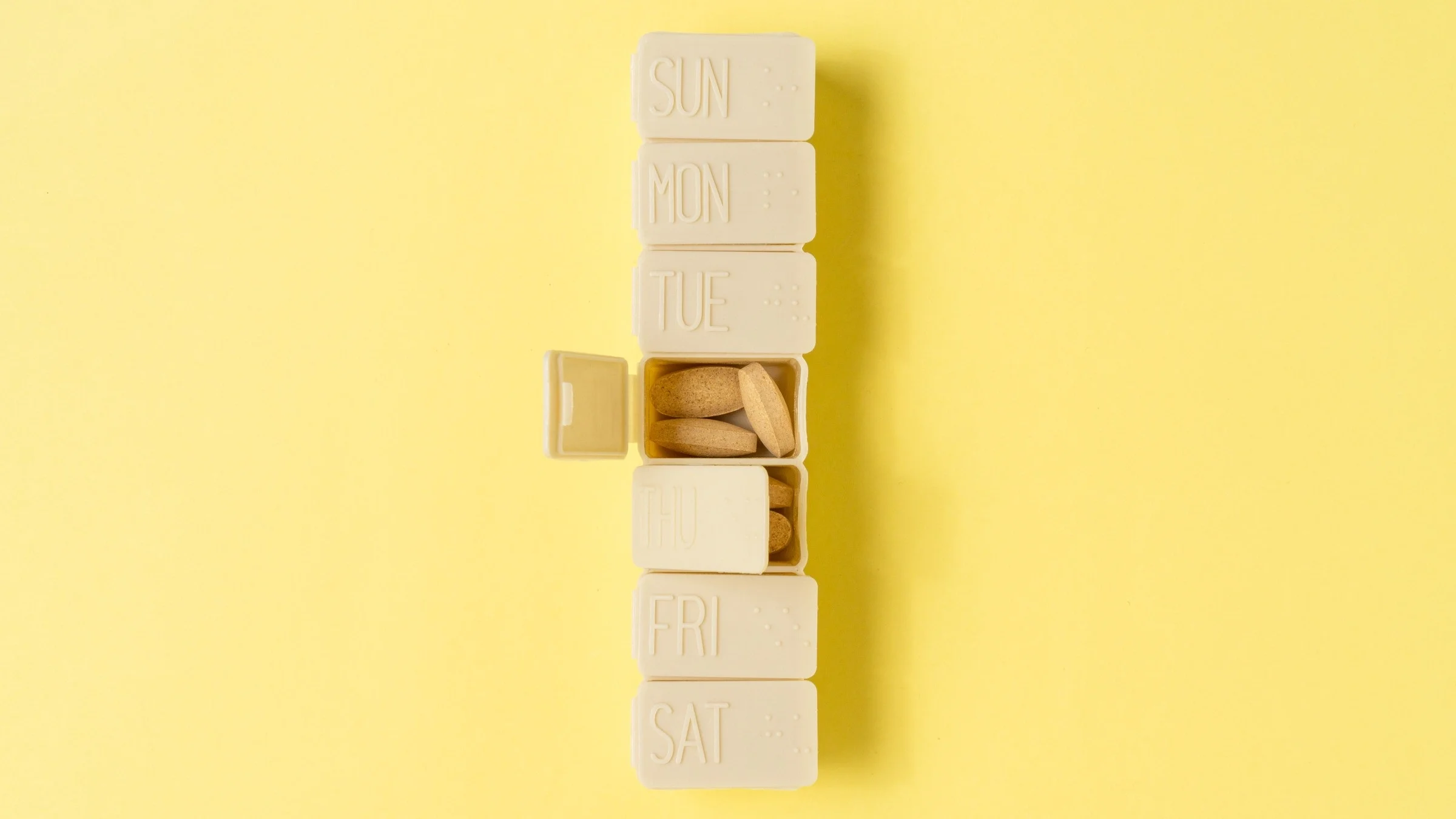 Tan pill organizer on a light yellow background one of the days of the week is open to show vitamins inside.