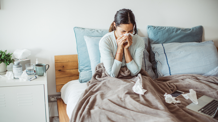 A woman blowing her nose in bed. An assortment of tissues, mug, glass of water, and medications are on her bedside table. 