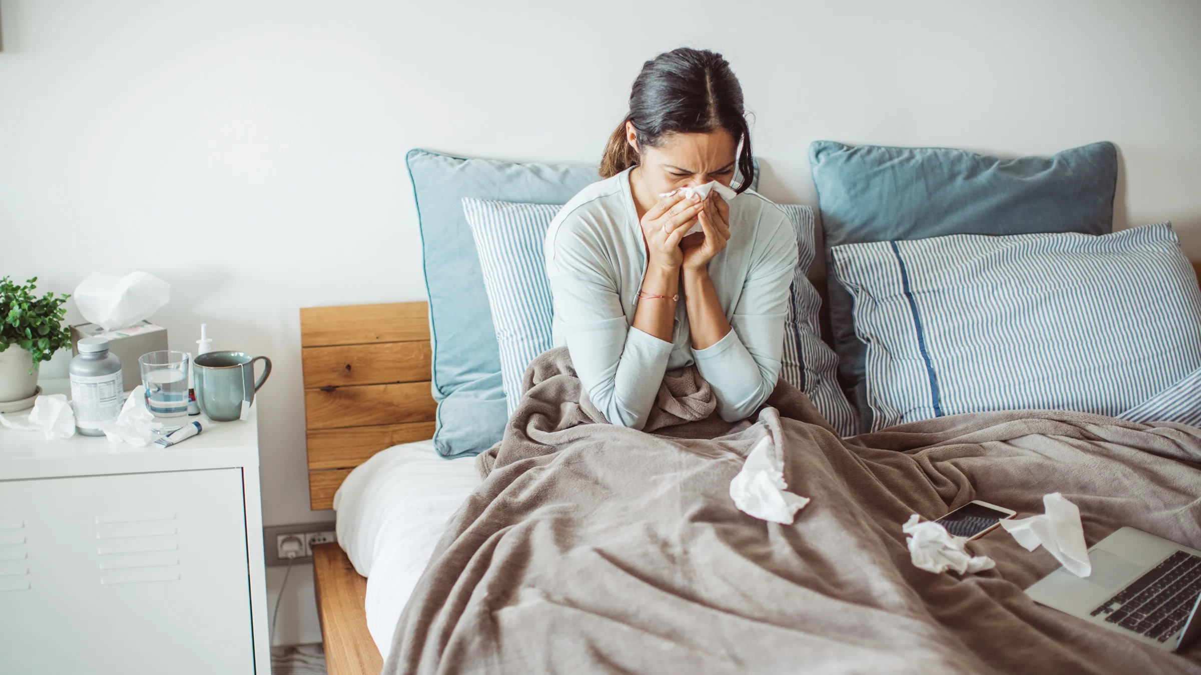 A woman blowing her nose in bed. An assortment of tissues, mug, glass of water, and medications are on her bedside table.