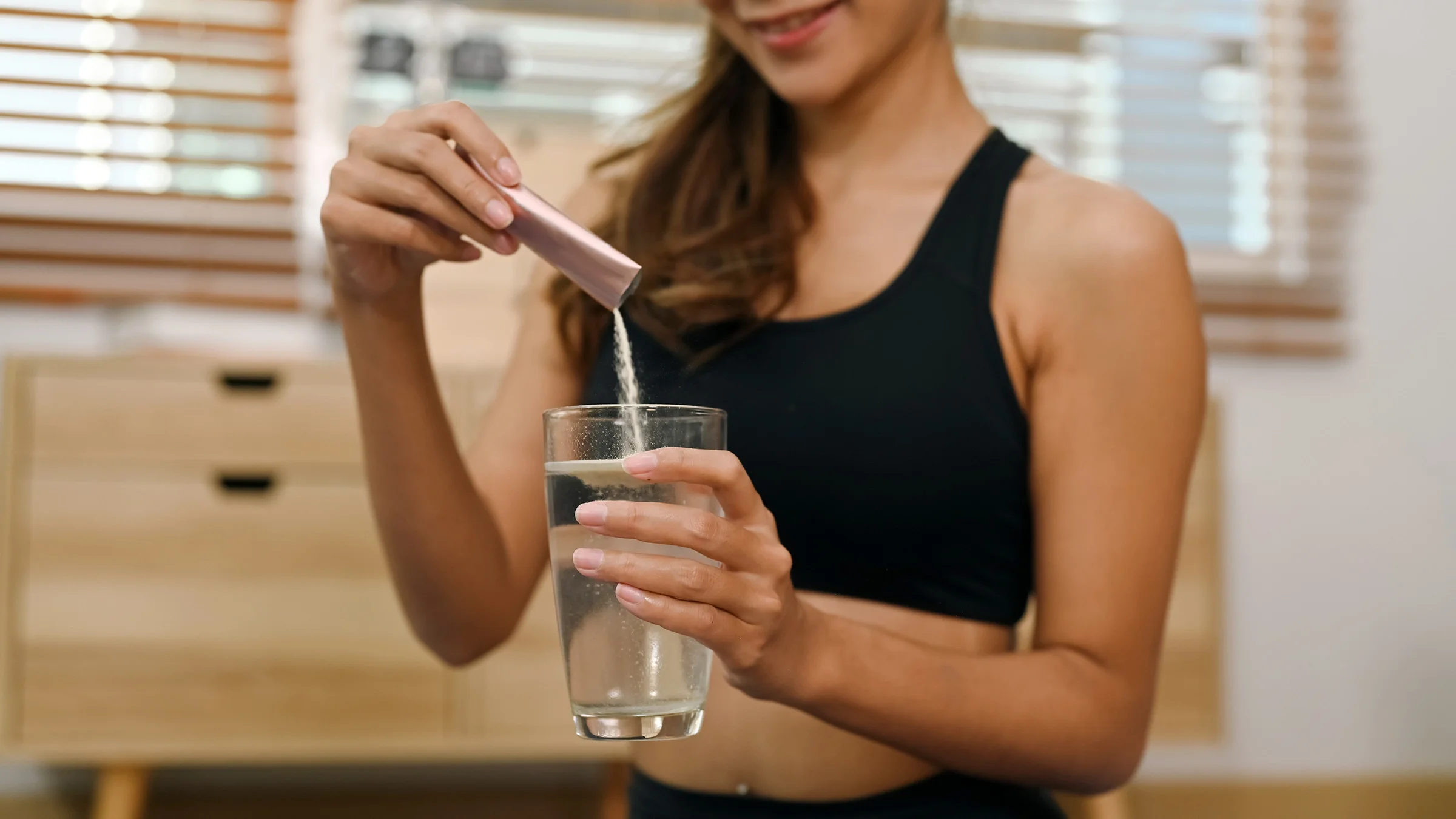 A woman pours a packet of collagen powder into a glass of water.