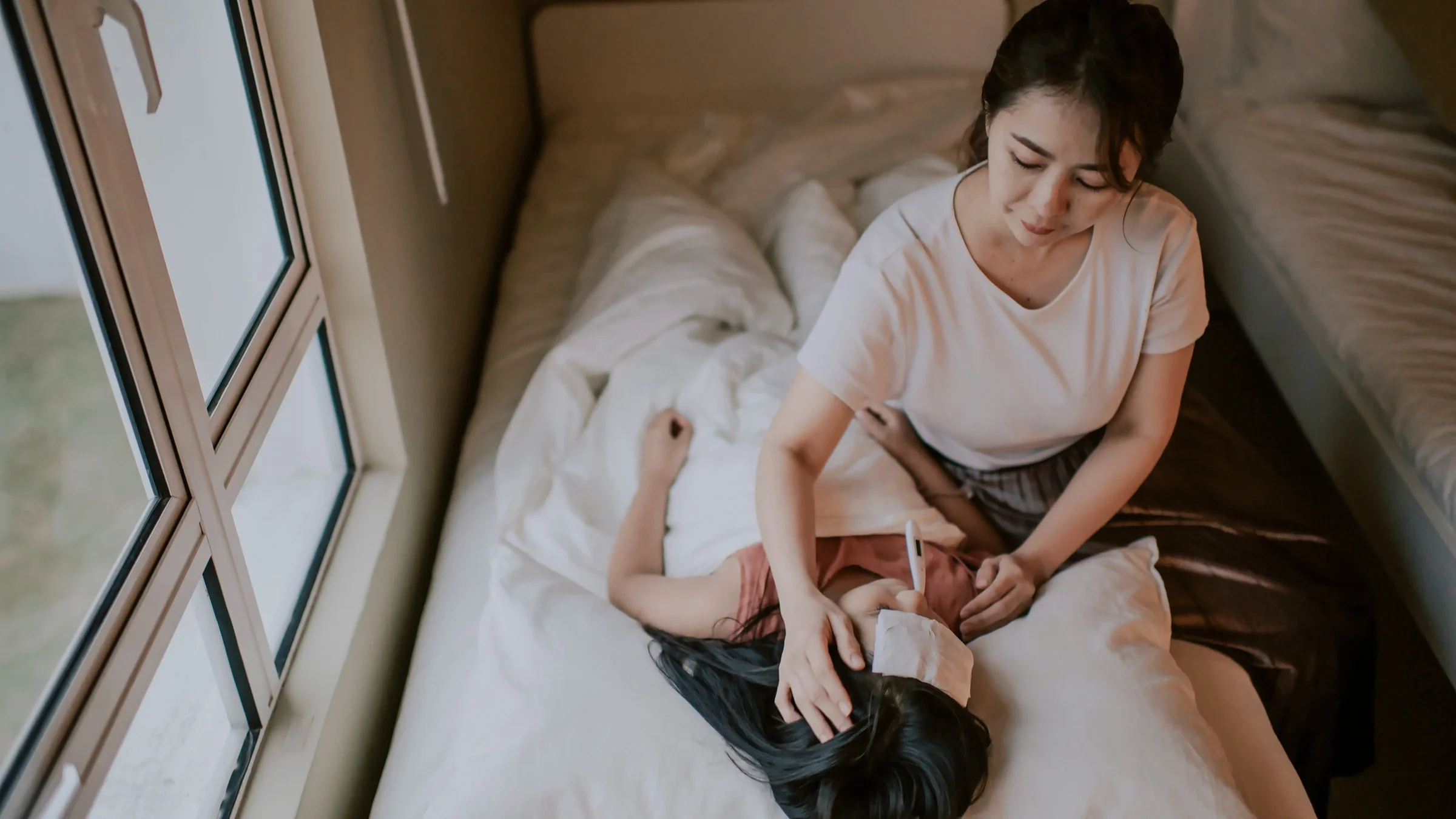 Mother taking her daughter's temperature and putting a compress on her forehead while she lies in bed.