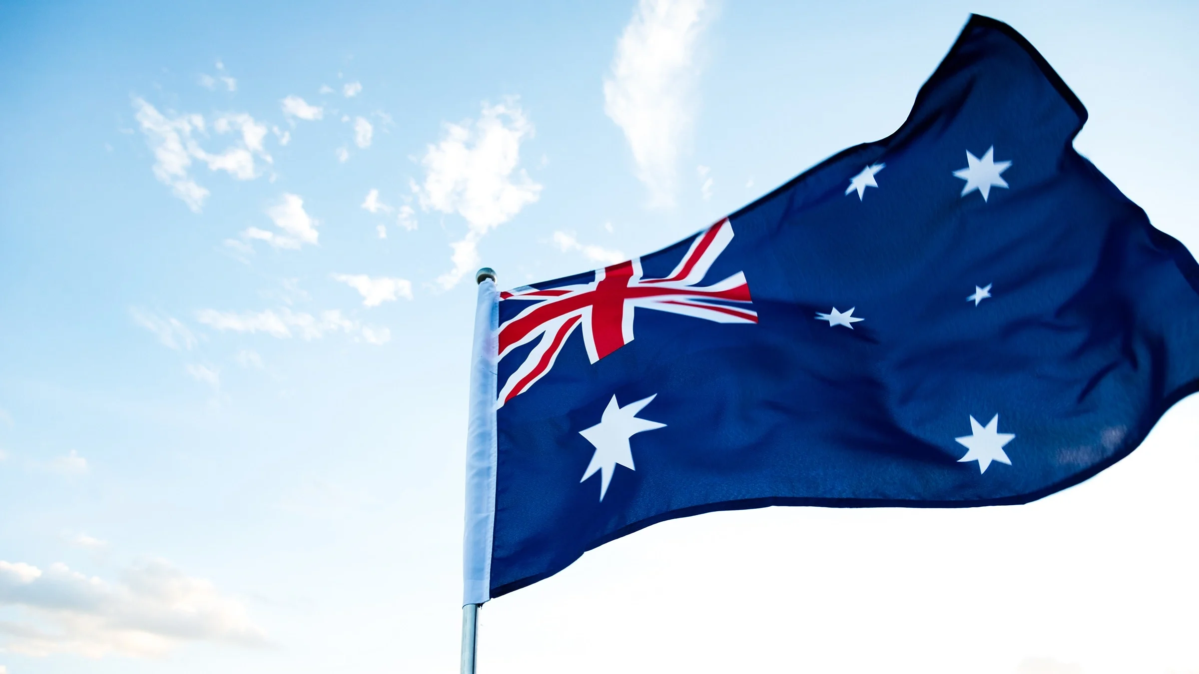 Australian flag flying on a clear blue sky day with some clouds behind it.