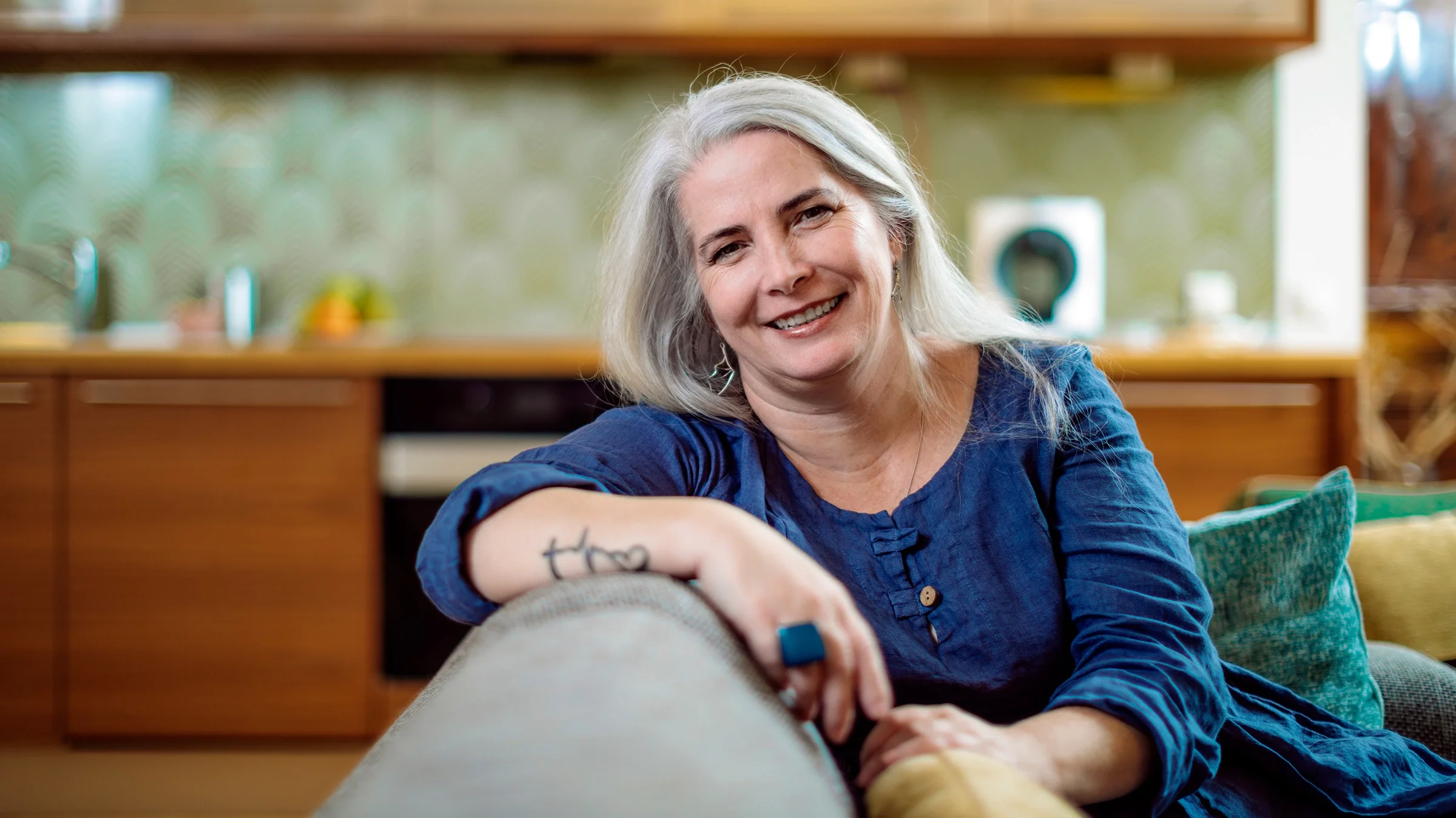 A woman smiles as she sits on her couch at home.