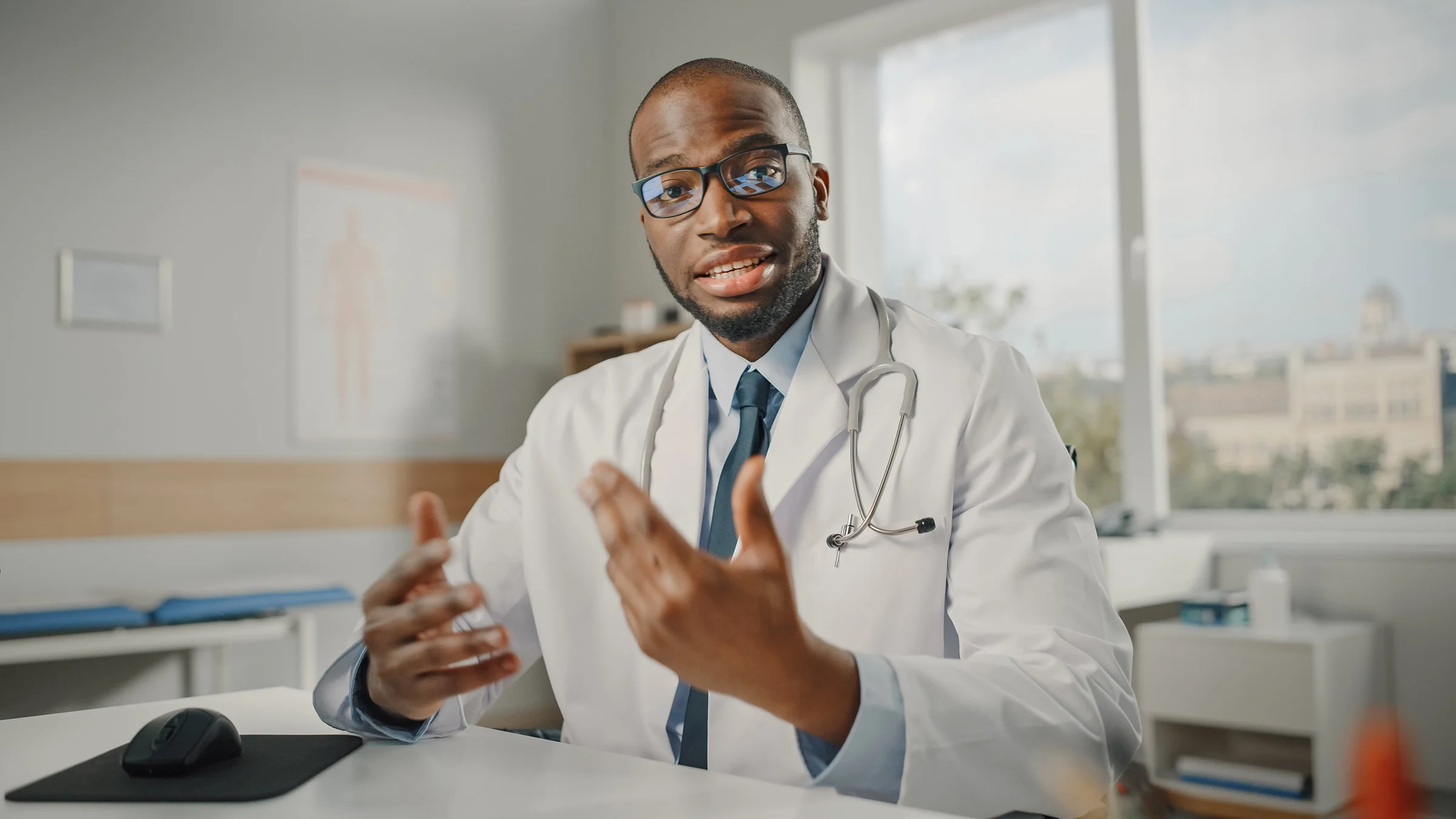 Doctor in a white coat and stethoscope talking directly to the camera. He is using his hands in an expressive way.