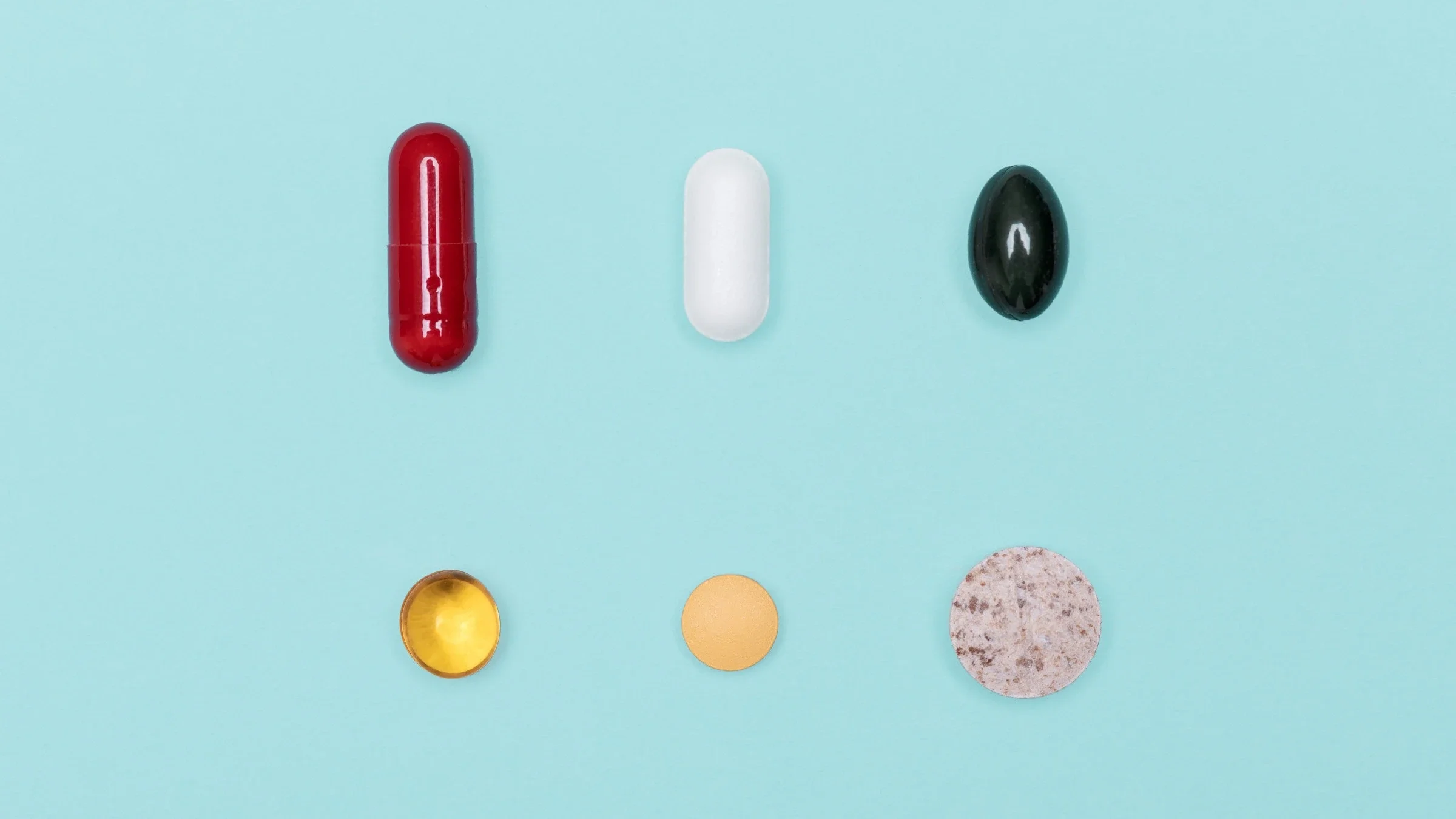 Six different supplement pills of various shapes, sizes, and colors laid flat on a light blue background.