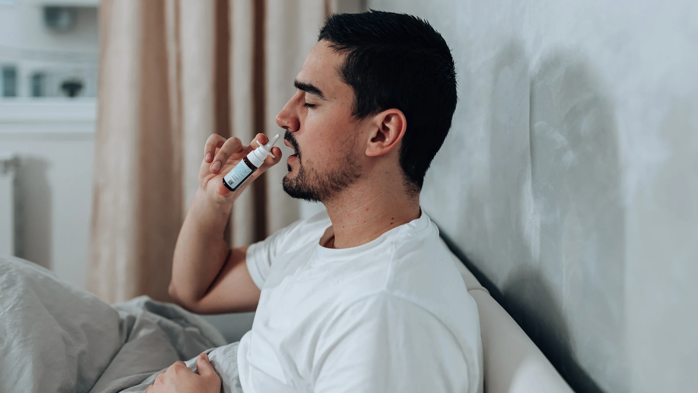 A man uses a nasal spray while sitting on his bed. 
RealPeopleGroup/iStock via Getty Images Plus     
