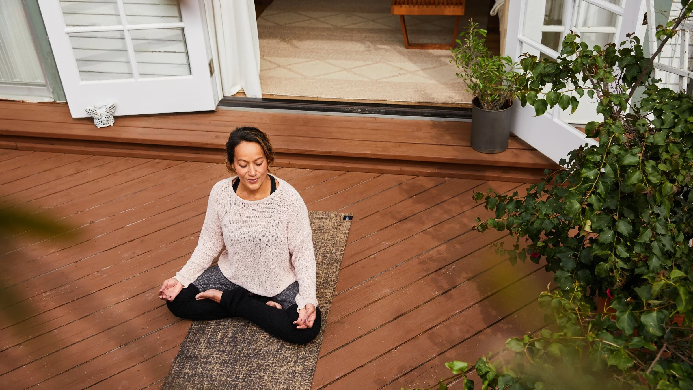 Over head shot of a woman sitting in lotus pose on her yoga mat on her outdoor deck with french doors open on the house.