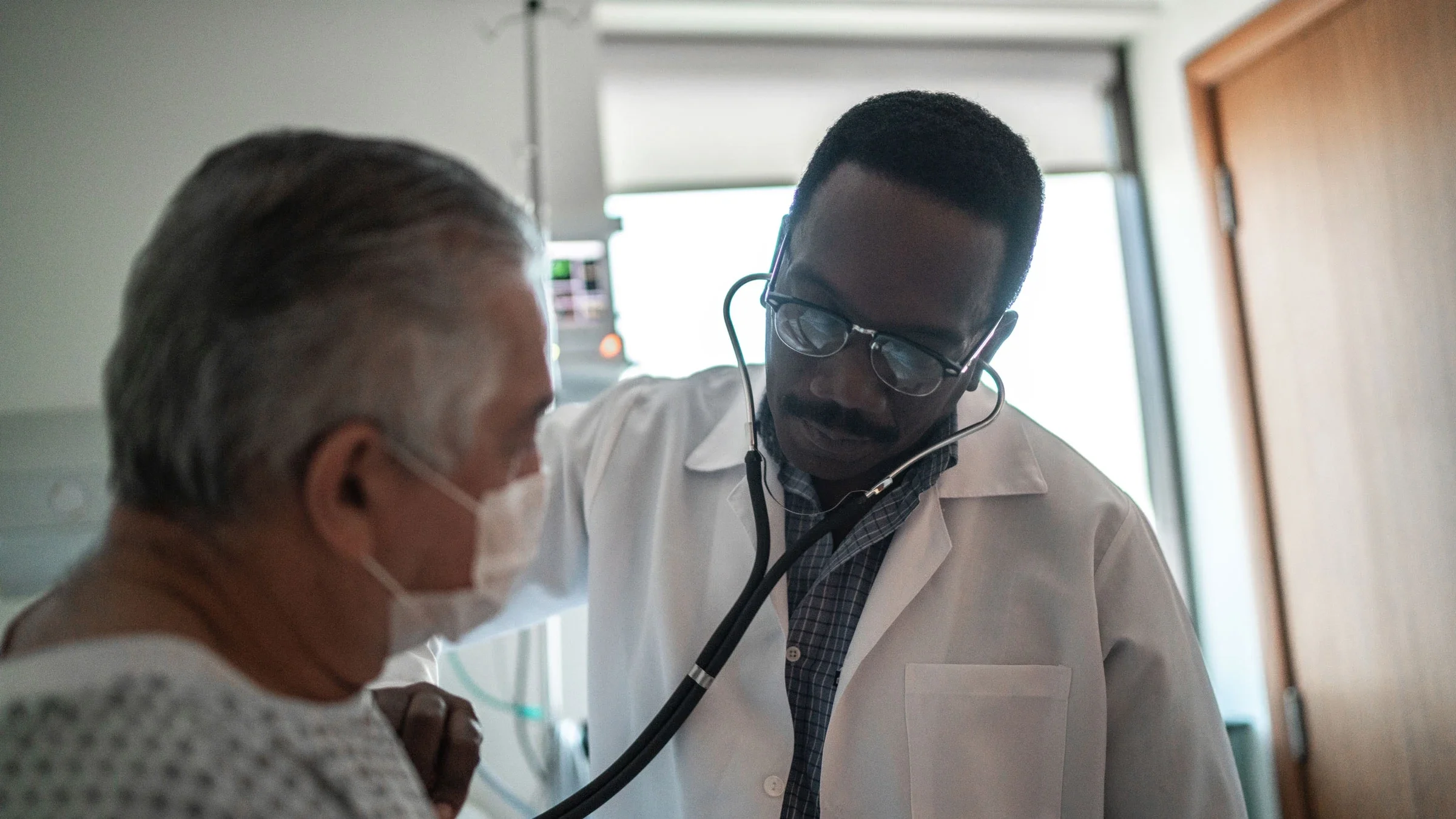 A healthcare provider is listening to an adult’s heartbeat in a hospital room.
