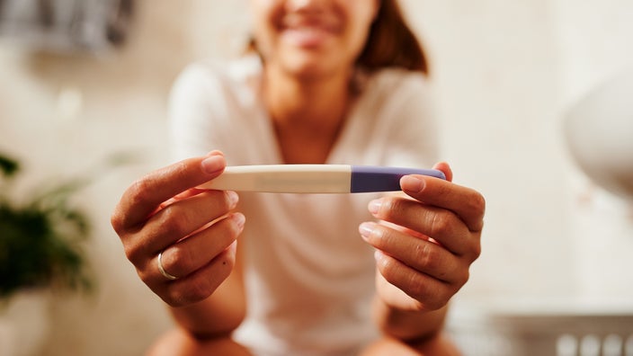 Cropped shot of a woman holding a pregnancy test in her hands. She is sitting in the bathroom and you can see her slight smile blurry in the background.