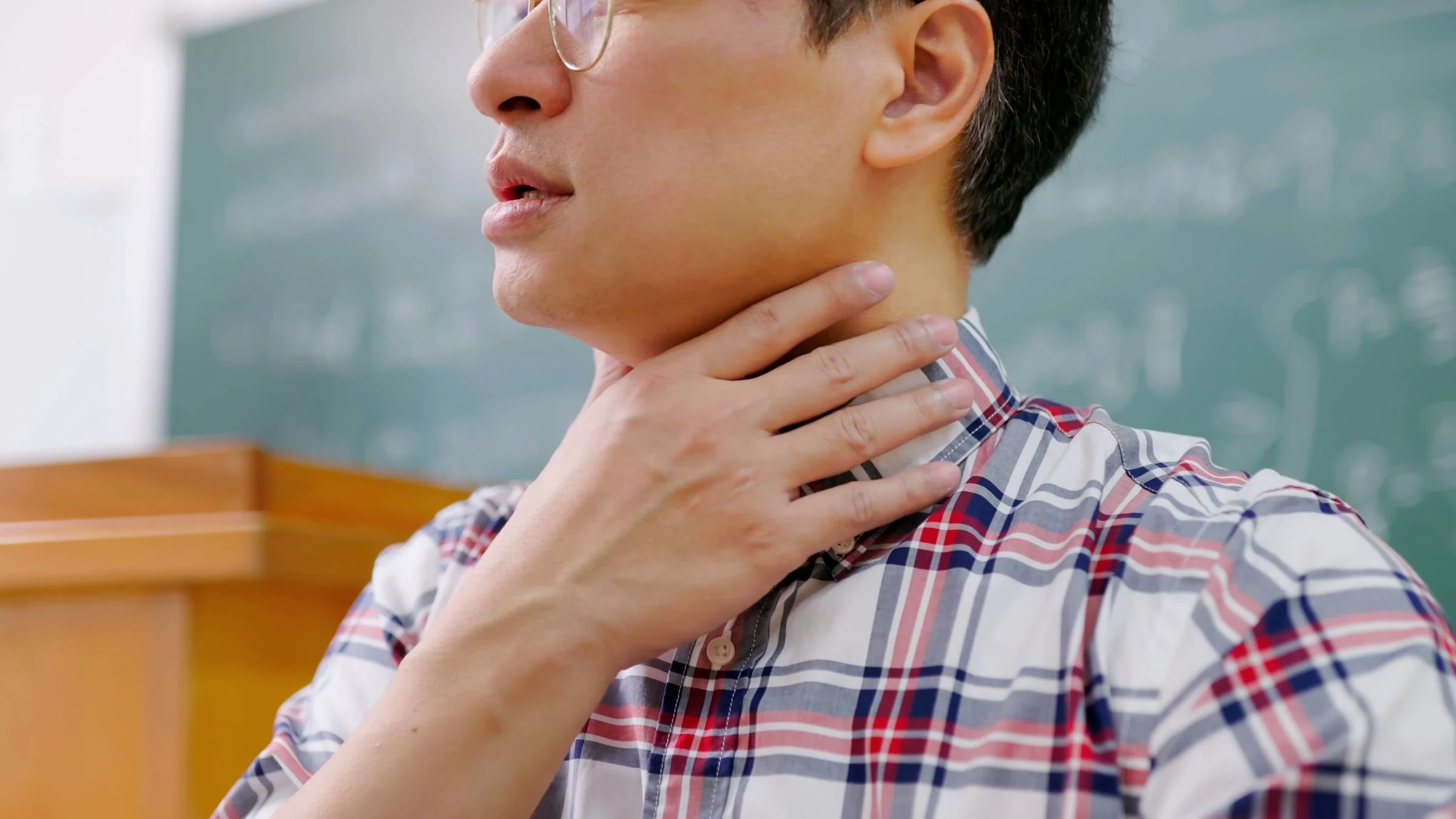 A close-up of a professor touching their vocal cord area, while trying to speak to their class.