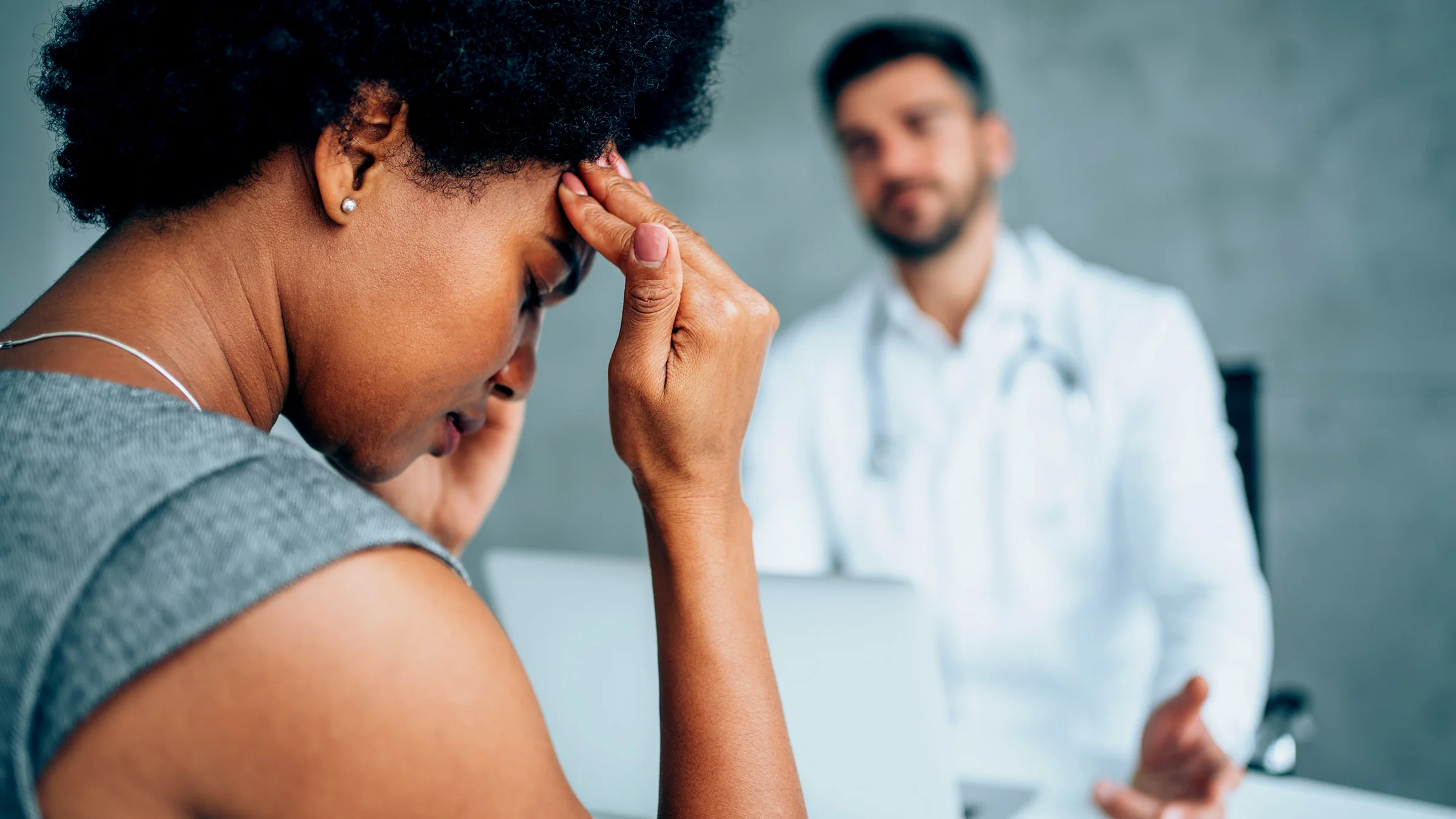 Black woman frustrated when her doctor doesn't believe her. The doctor is blurry in the background and has a judgemental attitude.